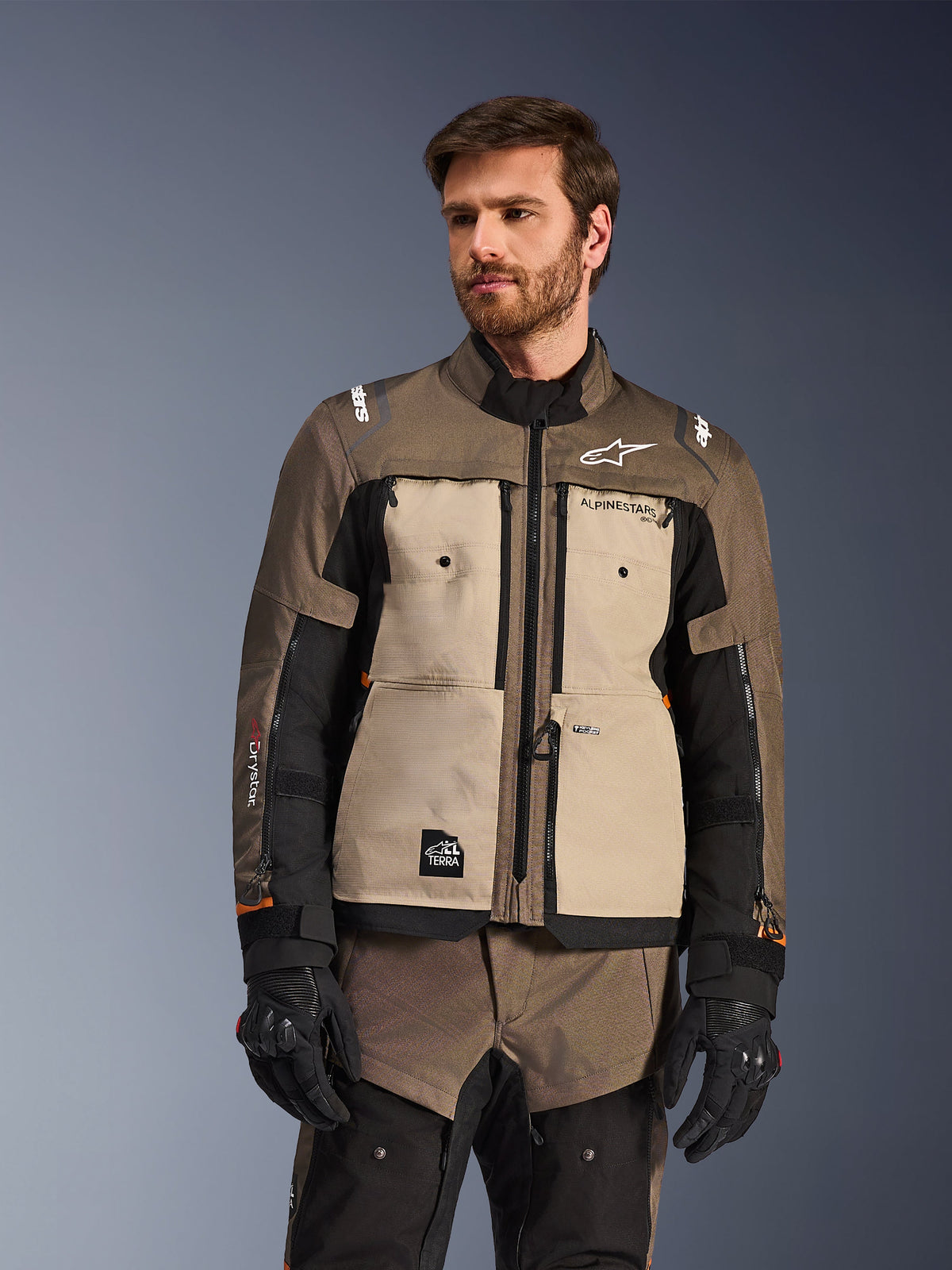 Alpinestars Cusco Drystar® Jacke, Adventure-Motorradjacke, Walnut Aluminum Black, von einem Modell mit schwarzen Handschuhen getragen, mit hellbraunen Brustpaneelen und schwarzen Ärmeln mit Alpinestars-Branding und All-Terra-Patch, schützende Allwetter-Tourenbekleidung