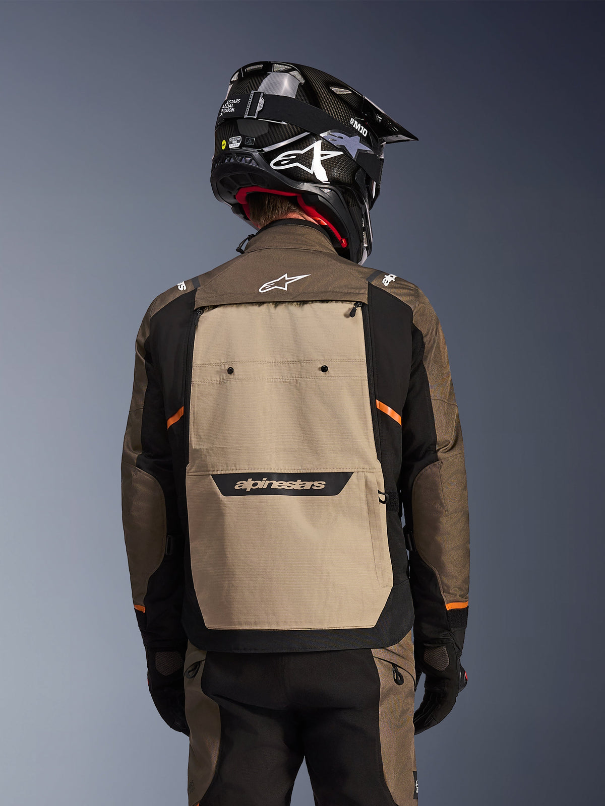 Rückansicht der Alpinestars Cusco Drystar® Jacke in Walnut Aluminum Black, Adventure-Touring-Motorradjacke mit einem großen sandfarbenen Rückenteil und Alpinestars-Branding, schwarze Ärmel mit orangefarbenen Akzenten, abgebildet mit einem schwarzen Carbon-Helm.