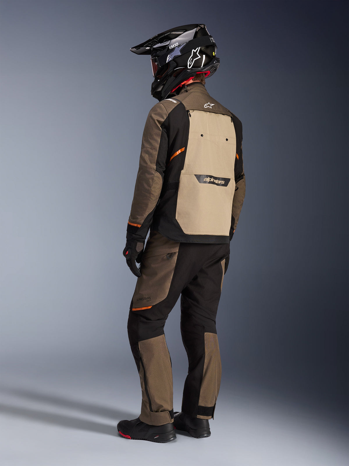 Alpinestars Cusco Drystar® Jacke, Adventure-Tourenjacke, Walnut Aluminum Black, Rückansicht mit beigem Rückenteil und orangefarbenen Akzenten, getragen mit passender Hose und schwarzem Carbon-Helm.