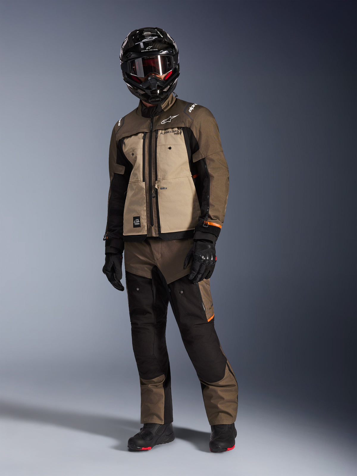 Alpinestars Cusco Drystar® Jacke, Walnut Aluminum Black, getragen von einem Fahrer in kompletter Adventure-Touring-Ausrüstung, sandfarbene und braune Einsätze mit schwarzen Akzenten, passend kombinierte Hose, schwarzer Integralhelm mit Schutzbrille, schwarze Stiefel, Studiohintergrund
