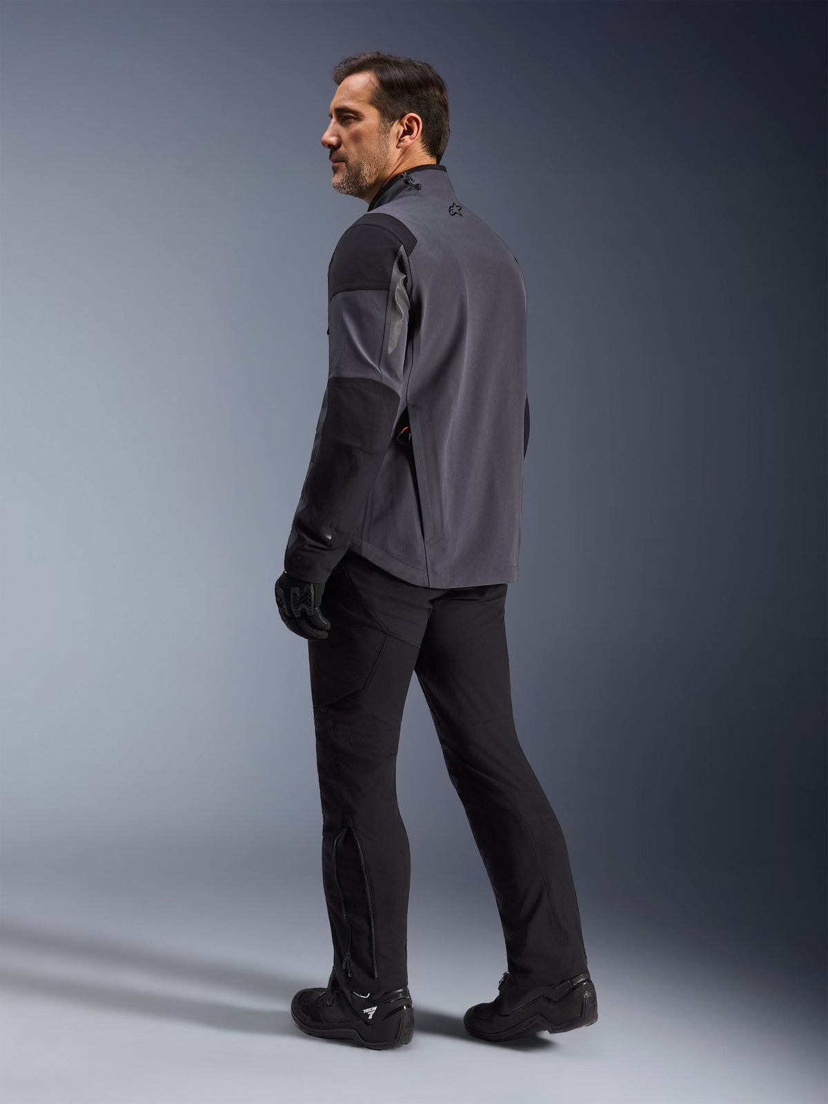 Alpinestars Maxdura Drystar®XF Laminierte Gepanzerte Jacke, Adventure-Motorradjacke, Dunkelgrau und Schwarz, Rückansicht eines Modells, das über die Schulter blickt, getragen mit schwarzer Motorradhose und Stiefeln, technisches 3-Lagen-Laminat-Design für Allwetter-Performance