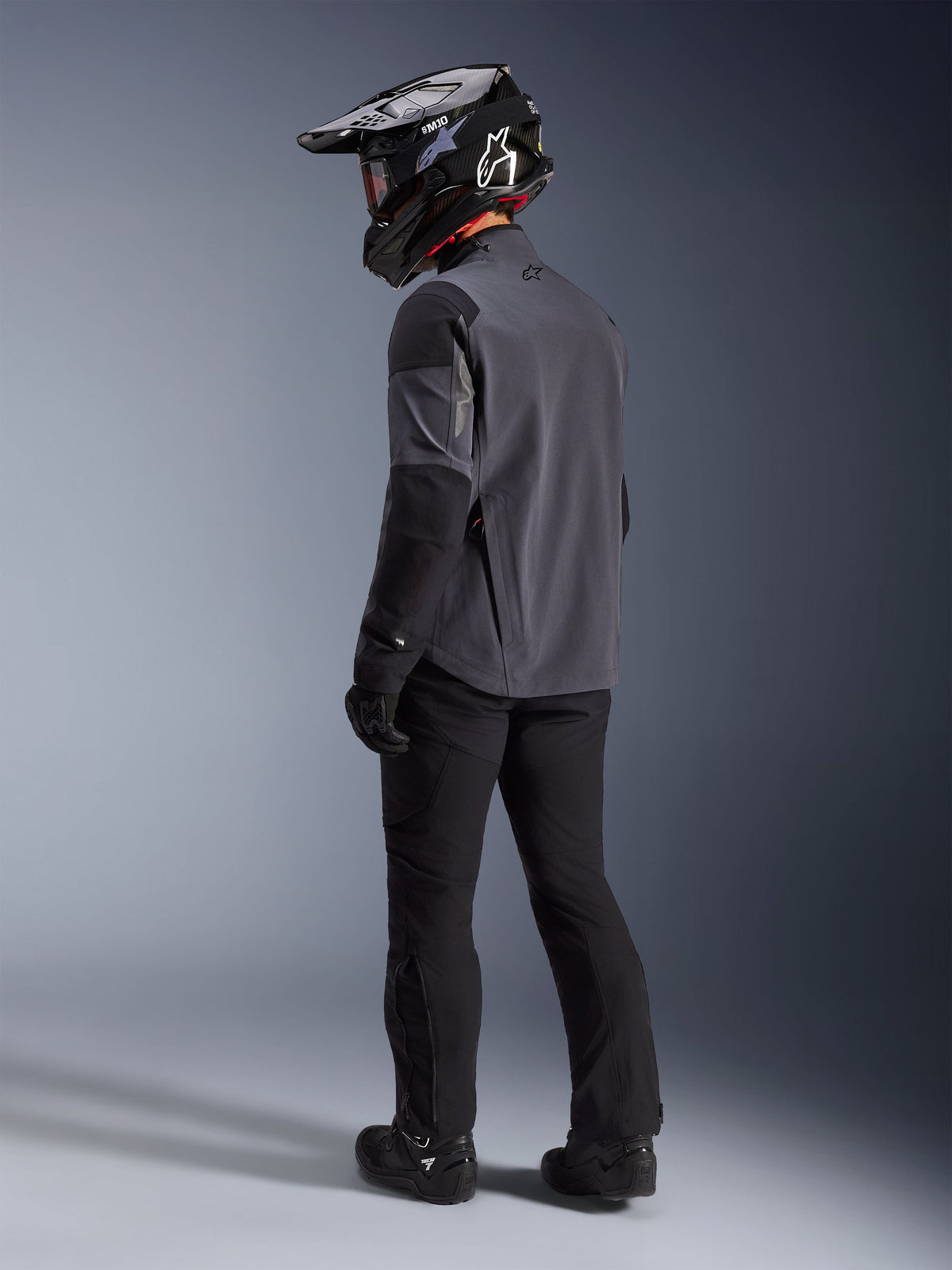 Adventure-Fahrer in der Maxdura Drystar®XF Laminierten Gepanzerten Schale in Deep Gray Black, Rückansicht. Komplette Ausrüstung inklusive schwarzer Motorradhose, einem schwarzen Motocross-Helm mit Alpinestars-Branding und schwarzen Offroad-Stiefeln. Technische Allwetter-Fahrausrüstung für Dual-Sport-Performance.