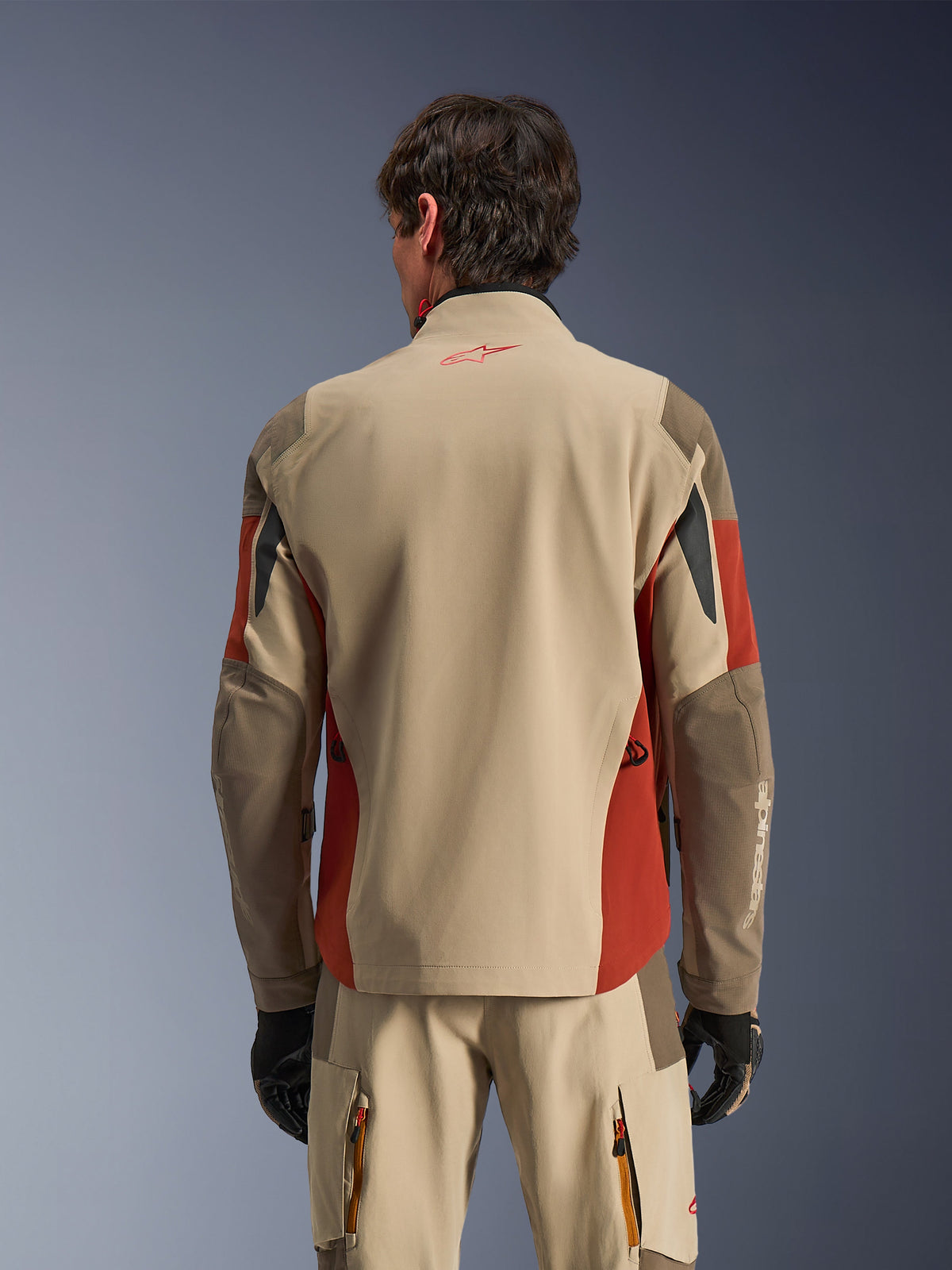 Man wearing Alpinestars Maxdura Drystar®XF Laminated Armored Shell from the back, Aluminum Walnut and Rust Brown adventure jacket, matching technical pants and black gloves, 3-layer laminated textile construction with reinforced panels, coordinated outfit for motorcycle touring