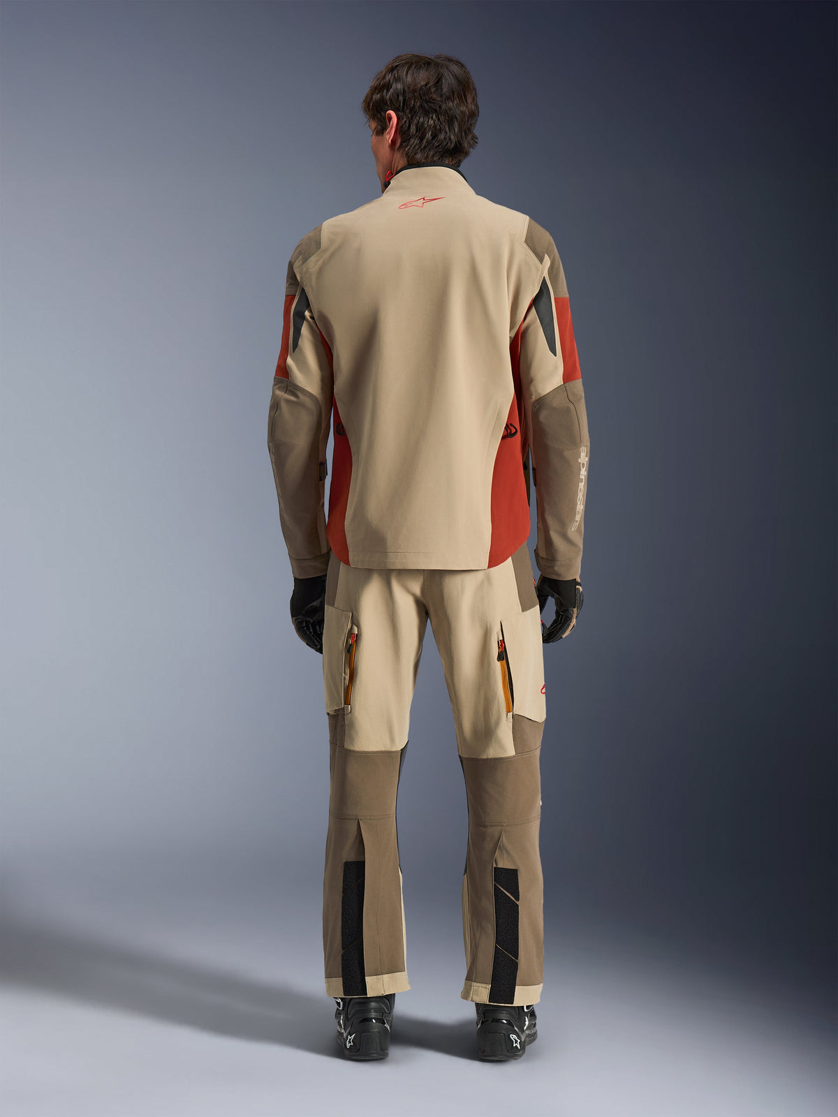 Rear view of a rider wearing the Alpinestars Maxdura Drystar®XF Laminated Armored Shell and matching adventure pants in Aluminum Walnut Rust Brown, tan and brown textile with orange accents, paired with black motorcycle boots and gloves, professional adventure touring gear.