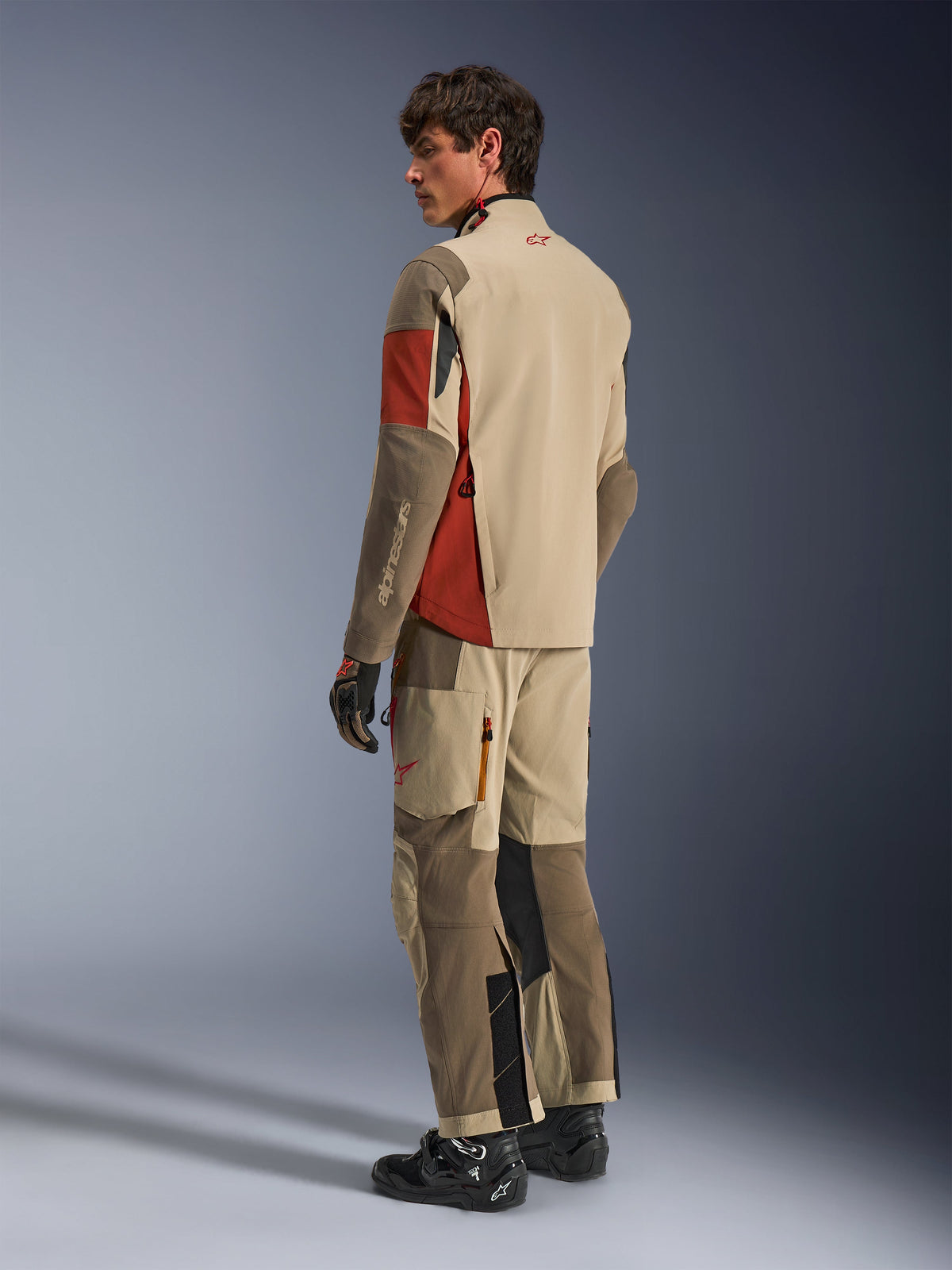 Male model wearing the Alpinestars Maxdura Drystar®XF Laminated Armored Shell in Aluminum Walnut Rust Brown, matching adventure touring pants, black gloves, and black boots, back view showing the technical textile construction and reinforced zones.