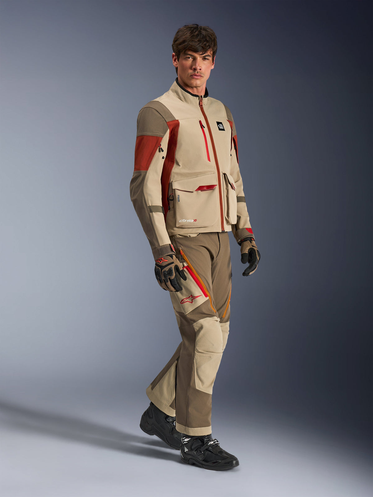 Male model wearing Alpinestars Maxdura Drystar®XF Laminated Armored Shell, Aluminum Walnut Rust Brown adventure jacket with matching pants, black and brown gloves, and black boots, featuring a multi-layered technical design with cargo pockets and red zipper details, coordinated outfit for adventure touring