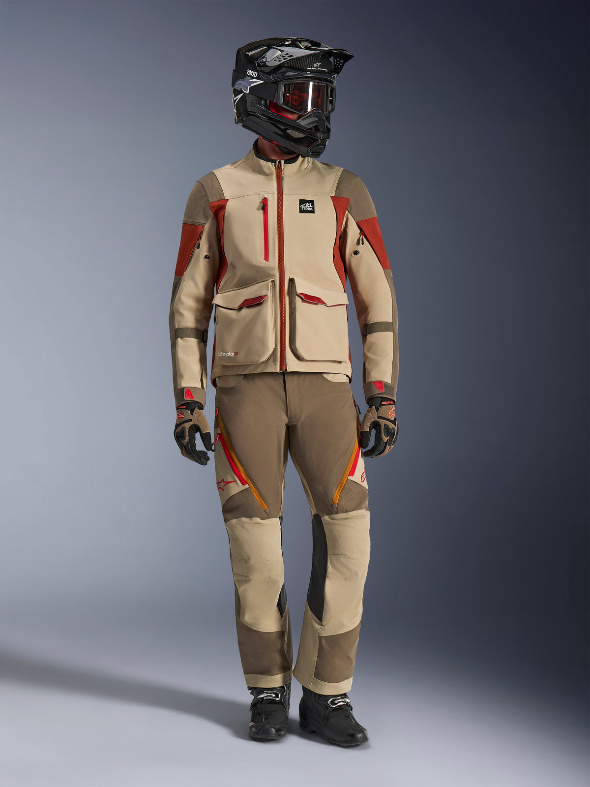 Full-body studio shot of a rider wearing the Alpinestars Maxdura Drystar®XF Laminated Armored Shell Jacket in Aluminum Walnut Rust Brown, paired with matching adventure pants, a black off-road helmet with red-tinted goggles, and technical gloves, showcasing a complete adventure touring outfit.