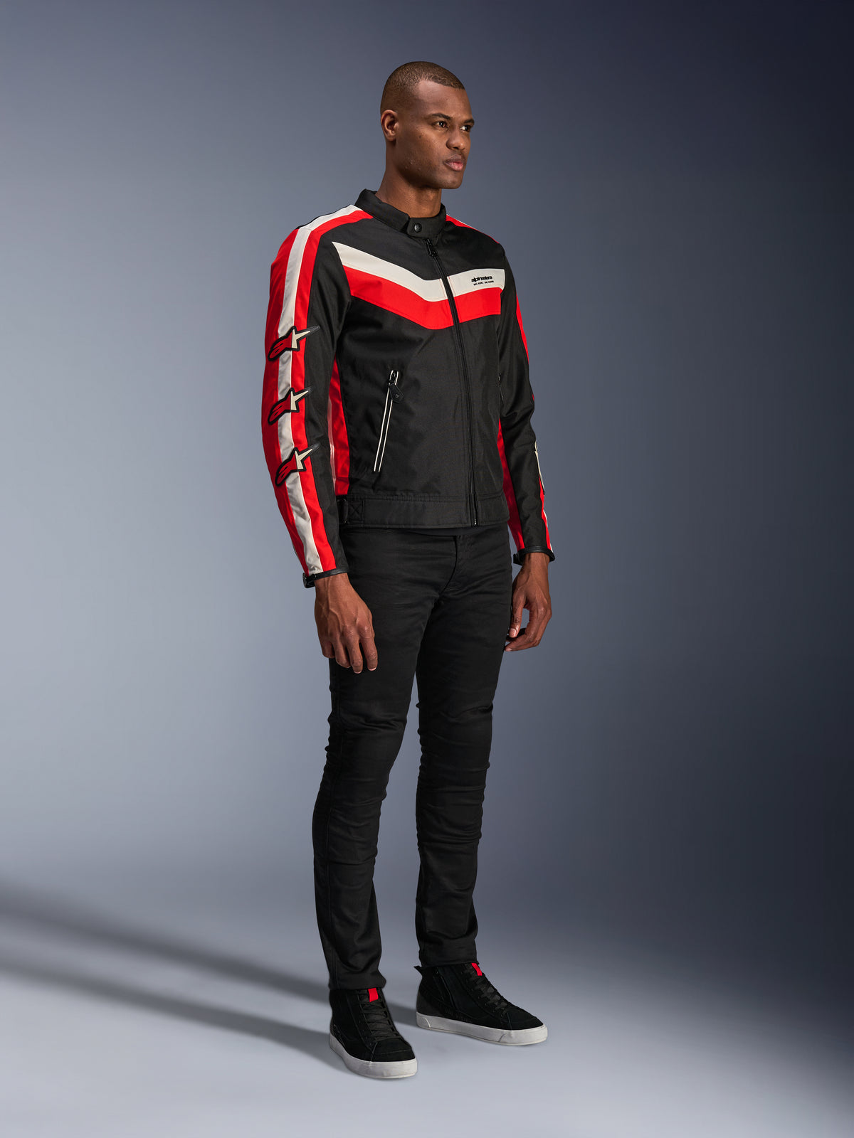 Männliches Model in der Alpinestars T-Dyno WR Jacke, Textil-Motorradjacke in Schwarz Bright Red Ecru, mit roten und cremefarbenen Streifen sowie Alpinestars Stern-Logos auf den Ärmeln, kombiniert mit einer schwarzen Hose und schwarzen High-Top-Sneakern, Vintage-inspirierte urbane Motorradausrüstung.
