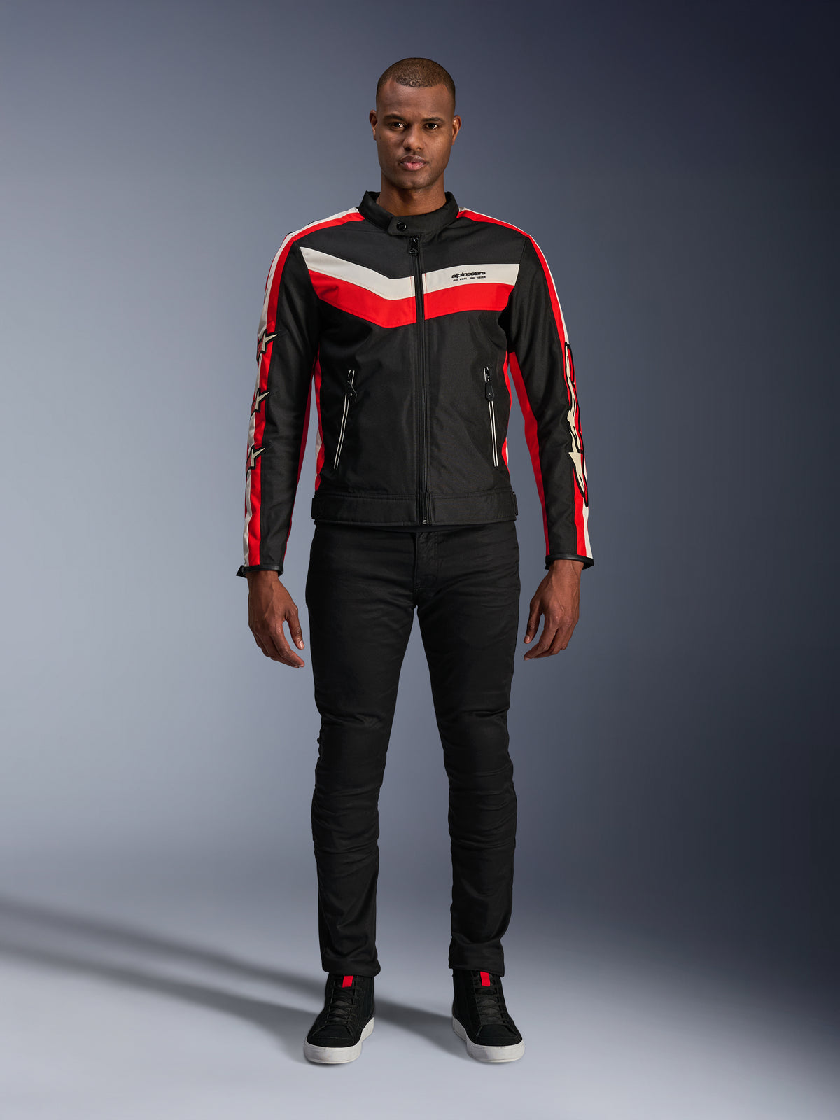 Alpinestars T-Dyno WR Jacke, Textil-Motorradjacke in Schwarz, Bright Red und Ecru, mit V-förmigen Bruststreifen und Stern-Branding an den Ärmeln, getragen mit schwarzer Hose und schwarzen Motorradschuhen, Heritage-inspirierte Urban-Ausrüstung
