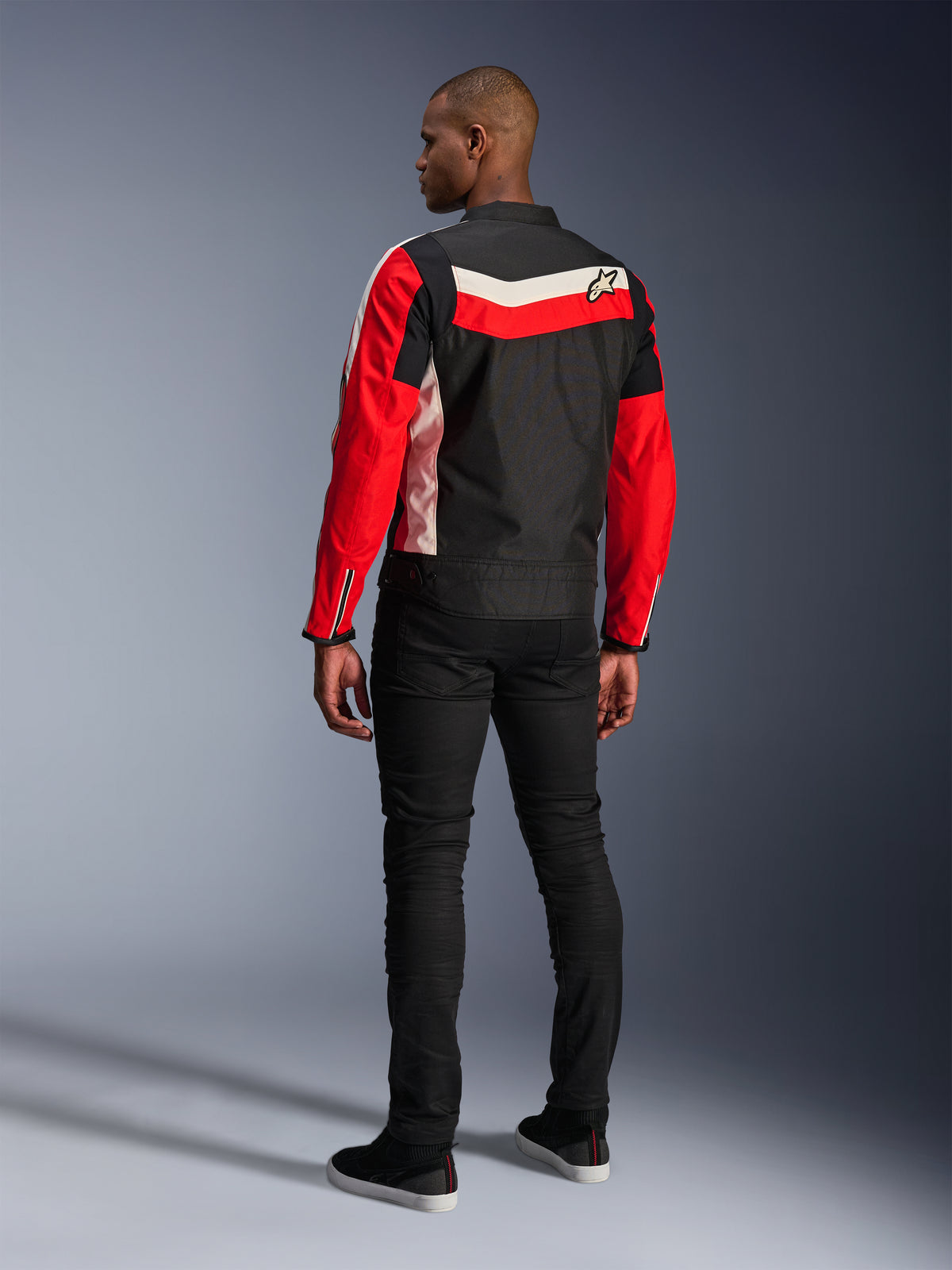 Alpinestars T-Dyno WR Jacke, Textil-Motorradjacke, Schwarz, Hellrot und Ecru, Rückansicht am Model, vintage-inspirierte Biker-Passform mit Heritage-Farbeinsätzen und Logo, moderne urbane Straßenausrüstung.