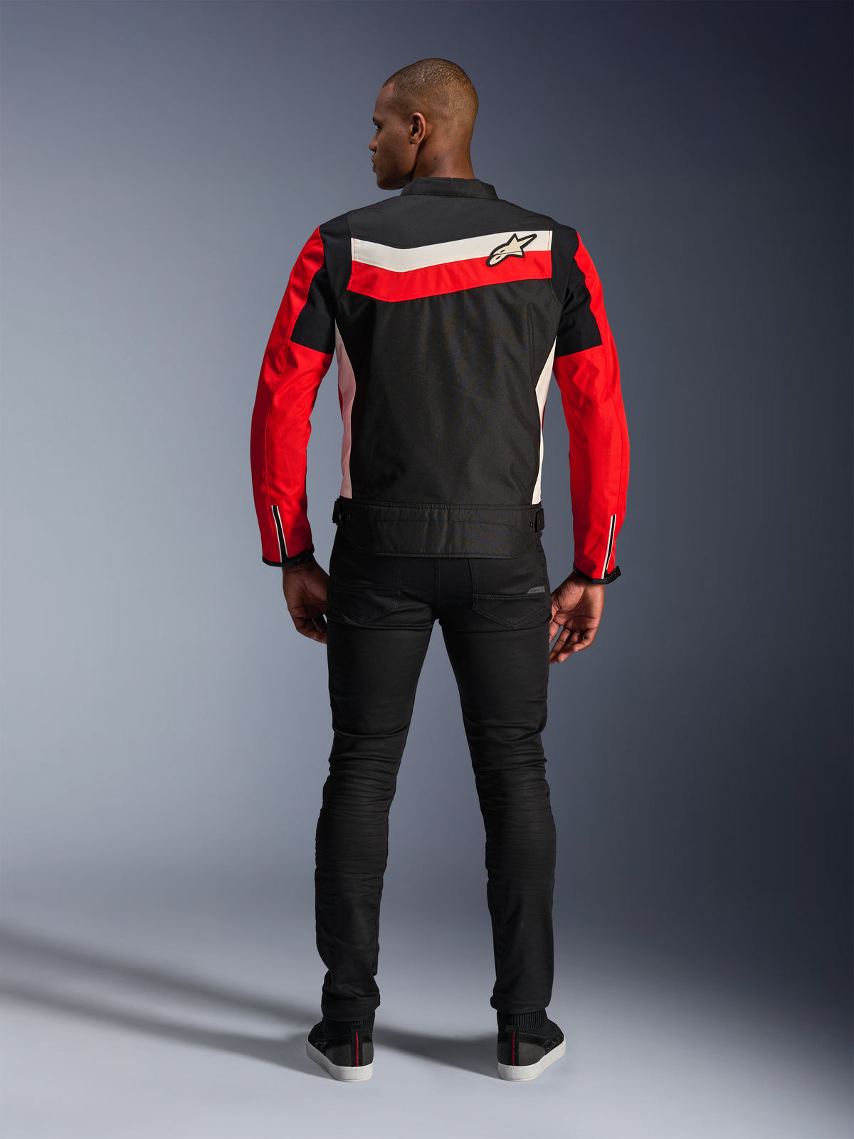 Alpinestars T-Dyno WR Jacke, Textil-Motorradjacke, Schwarz, Leuchtendes Rot und Ecru, Rückansicht mit horizontalen Rückenstreifen und roten Ärmeln, Vintage-inspirierte Biker-Passform, getragen mit schwarzer Hose, urbane Motorradbekleidung