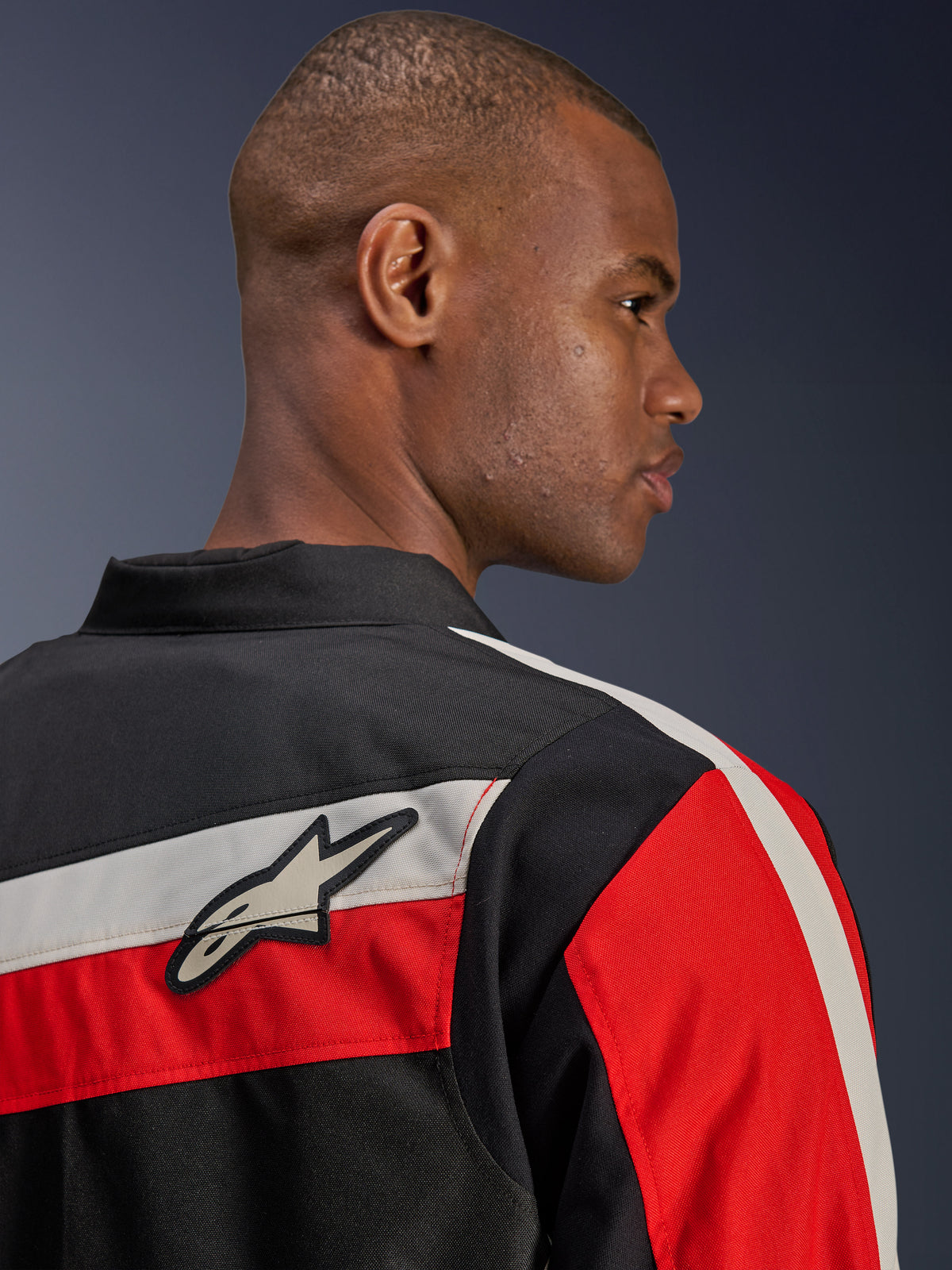 Alpinestars T-Dyno WR Jacke, Nahaufnahme der Rückansicht an einem Model, Textil-Motorradjacke in Schwarz mit horizontalen Streifen in Ecru und leuchtendem Rot, Alpinestars Stern-Logo-Patch auf der Schulter, Heritage-inspirierter Biker-Stil