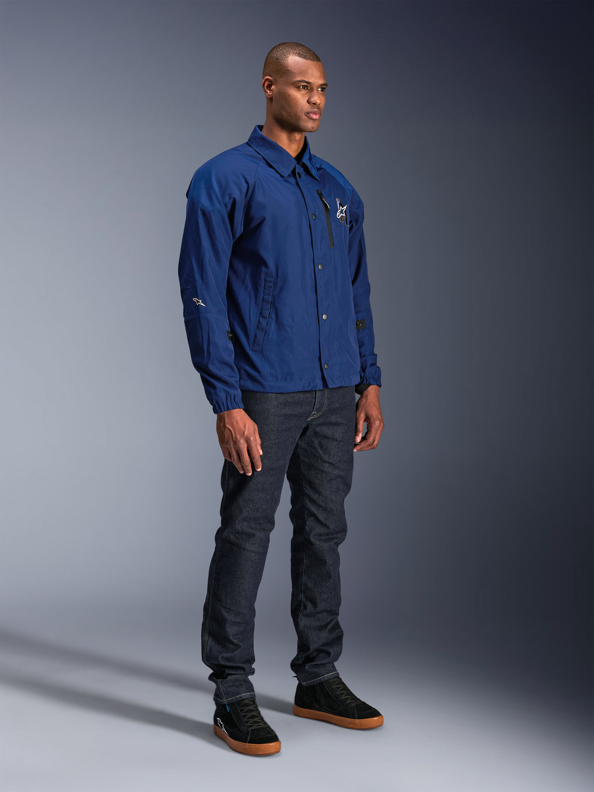 Alpinestars Revved WR Jacke, blaue Motorradjacke, getragen von einem männlichen Model mit dunkelblauen Jeans und schwarzen Sneakern, im wasserabweisenden Coach-Jacken-Stil mit Druckknöpfen vorne und minimalistischem Logo auf der Brust.