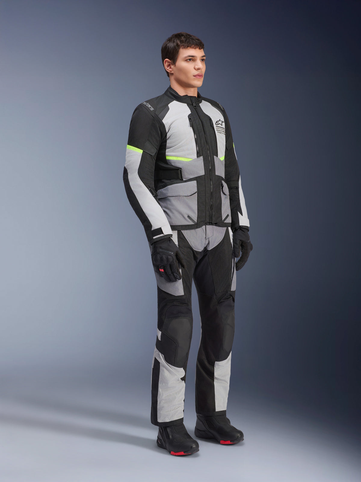 Alpinestars Andes Air Drystar® Jacke und Hose, Adventure-Touring-Motorradausrüstung, Eisgrau, Dunkelgrau und Schwarz mit neongelben Akzenten, getragen mit schwarzen Handschuhen und Stiefeln, Ganzkörper-Studioaufnahme für Touren in zwei Jahreszeiten.