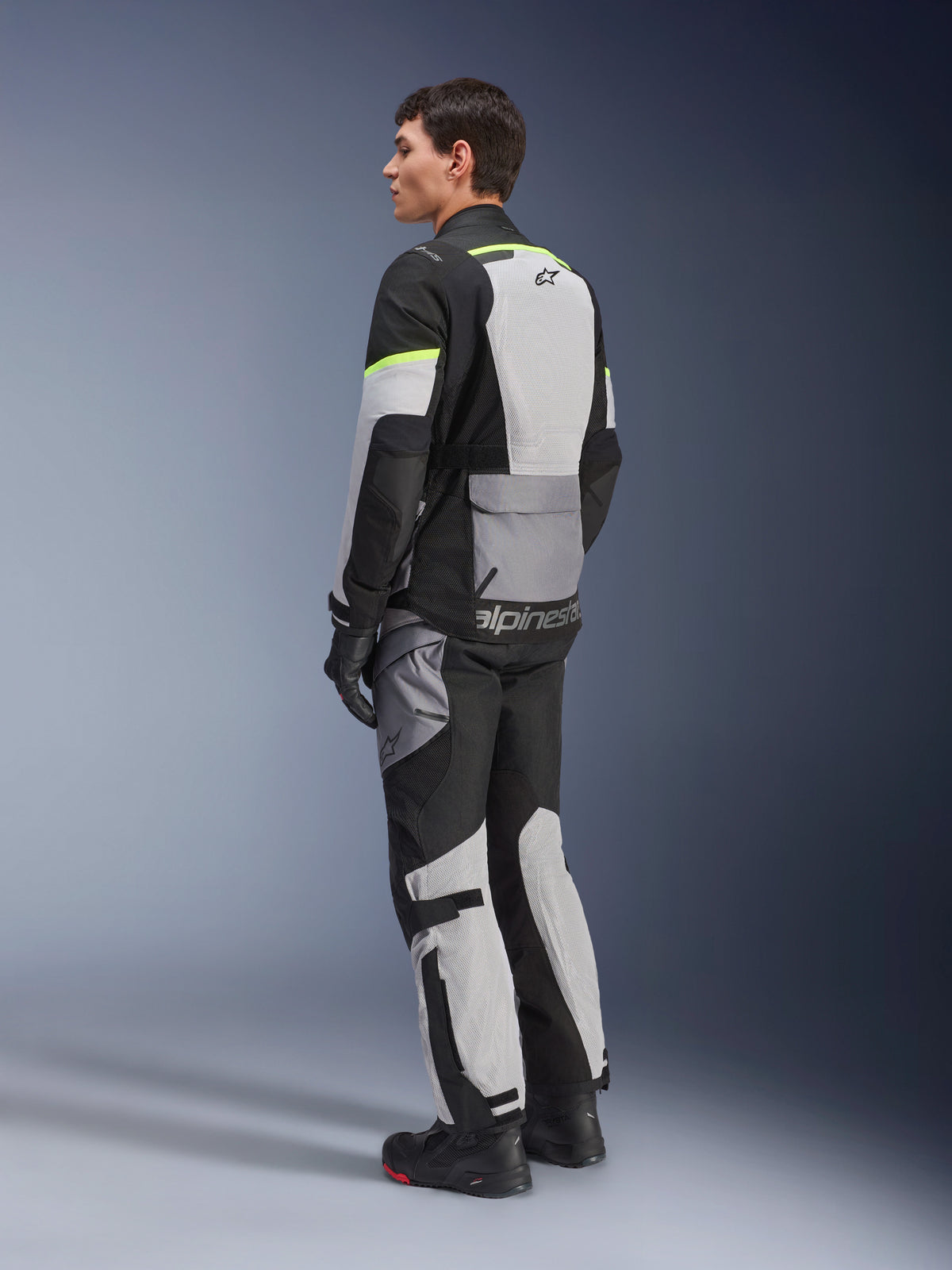 Fahrer trägt Alpinestars Andes Air Drystar® Jacke und Hose, Adventure-Tourenbekleidung in Ice Gray Dark Gray Black mit neongelben Akzenten, mit großflächigen Mesh-Einsätzen für Luftzirkulation, Alpinestars-Logo auf dem Rücken, kombiniert mit schwarzen Handschuhen und Stiefeln.