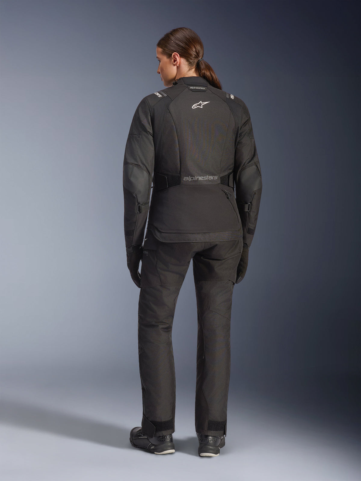 Motorradfahrerin in der Rückansicht mit der Alpinestars Stella Andes V4 Drystar® Jacke, Schwarz, kombiniert mit passender Motorradhose, weißes Astar-Logo auf dem Rücken, verlängerter Rückensaum, komplettes Adventure-Touring-Fahroutfit inklusive schwarzer Handschuhe und Stiefel