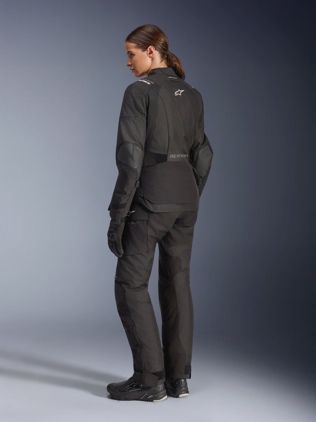 Motorradfahrerin in der Alpinestars Stella Andes V4 Drystar® Jacke, schwarze Adventure-Motorradjacke, Dreiviertel-Rückansicht, kombiniert mit passender Textilhose und Handschuhen, weiße Alpinestars-Logos am Rücken und an der Taille, verstärkte Schutzpaneele, verlängerter Saum am Rücken.