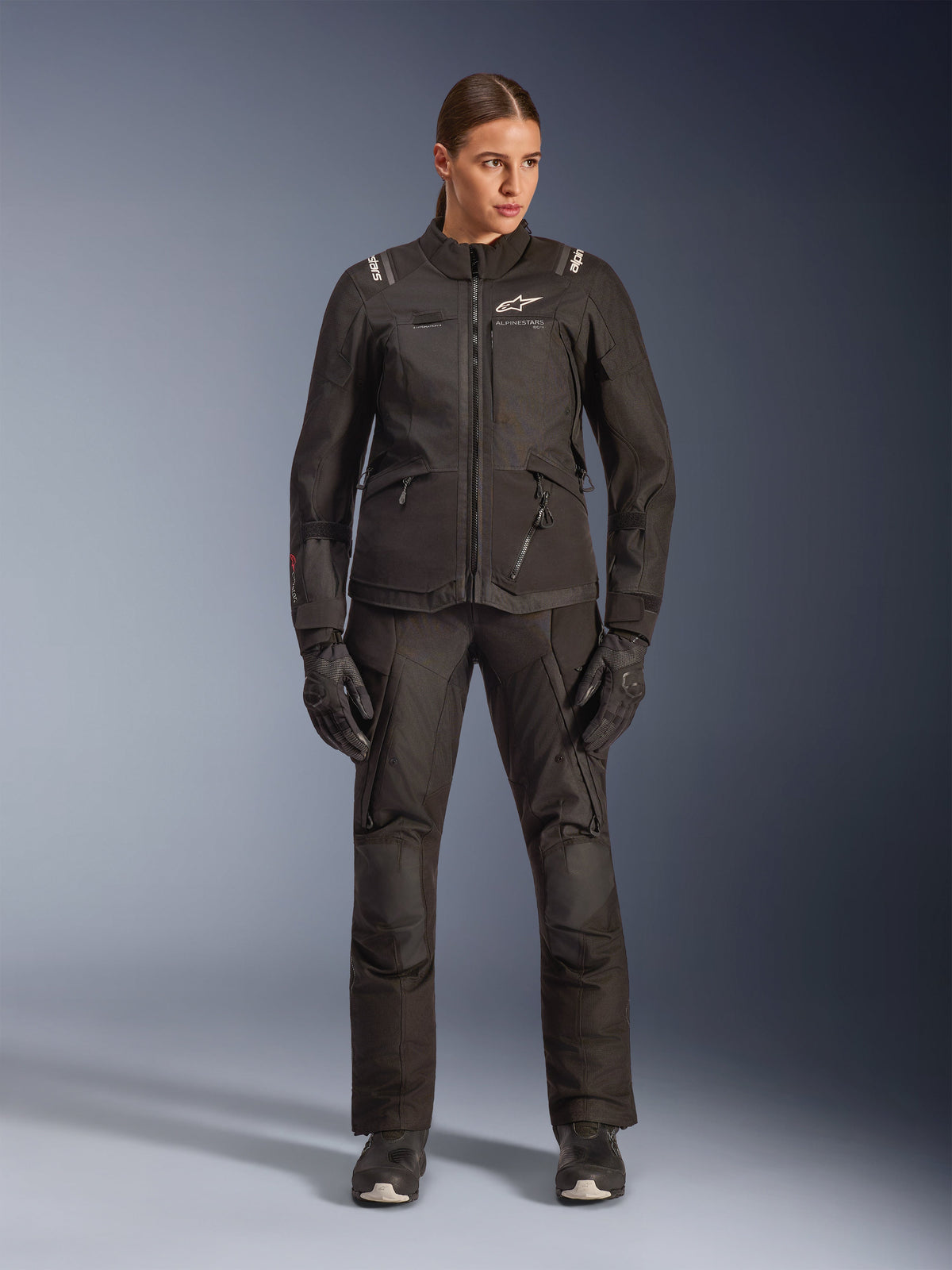 Motorradfahrerin mit Alpinestars Stella Andes V4 Drystar® Jacke in Schwarz, kombiniert mit passender schwarzer Adventure-Tourenhose und Handschuhen, technische Textilkonstruktion mit mehreren Funktionstaschen und weißem Alpinestars-Branding, komplette Allwetter-Motorradausrüstung