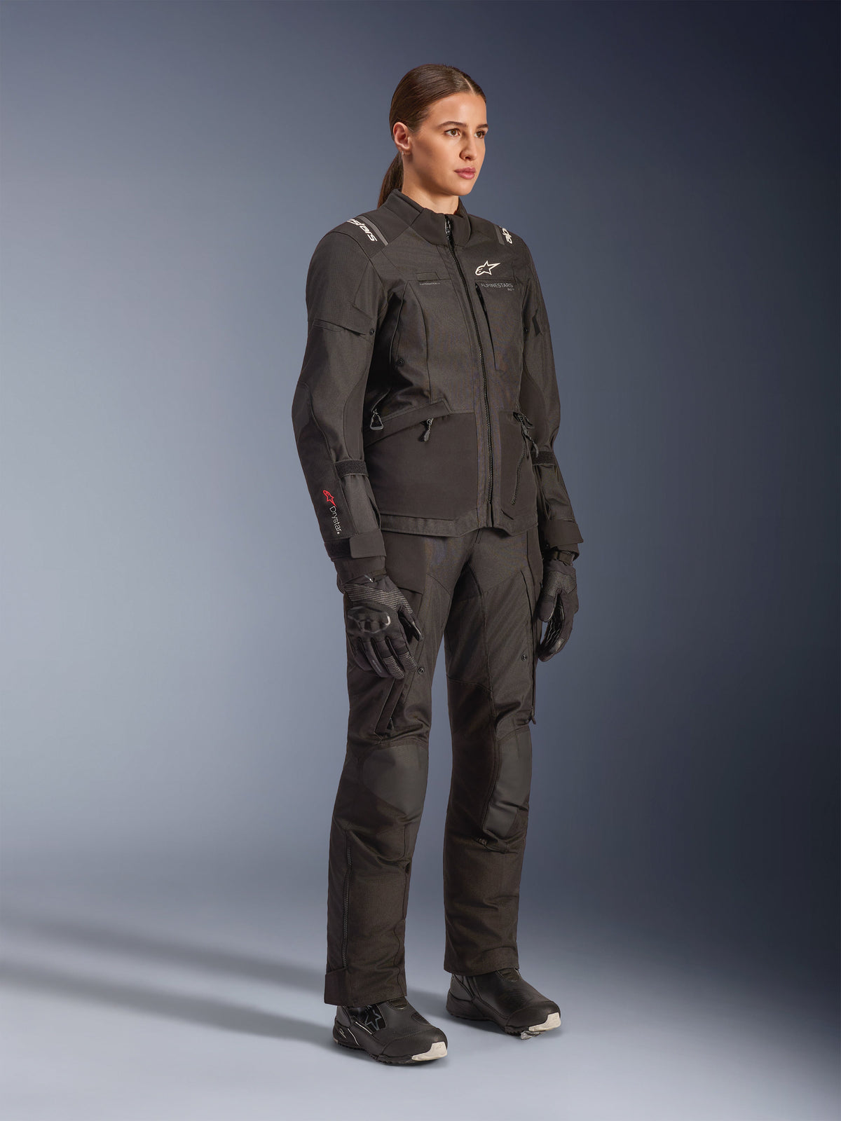 Motorradfahrerin in der schwarzen Alpinestars Stella Andes V4 Drystar® Damenjacke, kombiniert mit passender Adventure-Tourenhose, Handschuhen und Stiefeln, mit weißen Alpinestars-Logos und rotem Drystar-Branding, entwickelt für Motorrad-Performance bei jedem Wetter.