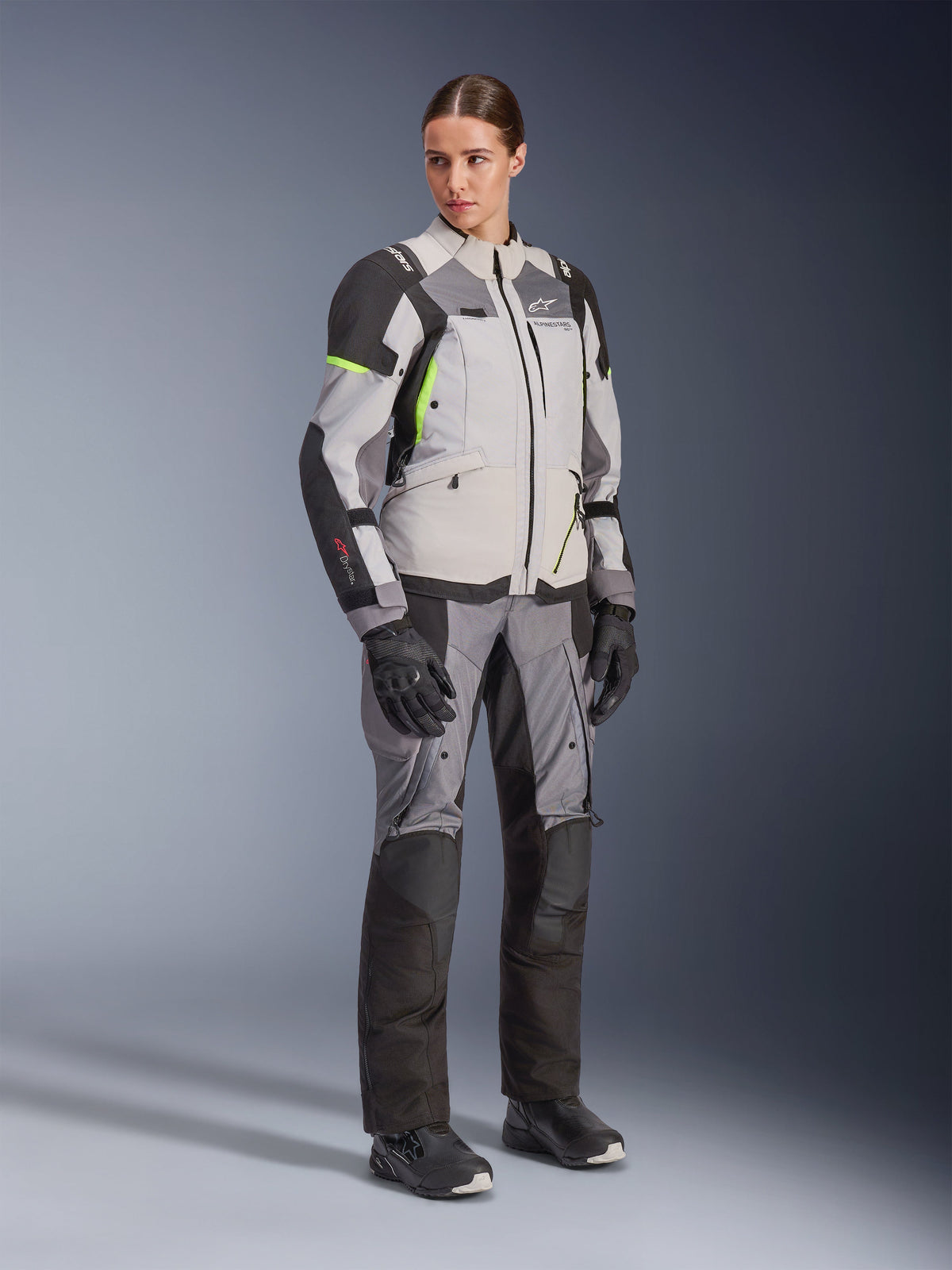Woman wearing Alpinestars Stella Andes V4 Drystar® Jacket and matching pants, Ice Gray and Dark Gray with Yellow Fluo accents, complete adventure touring gear set with black gloves and boots, full-length studio shot.