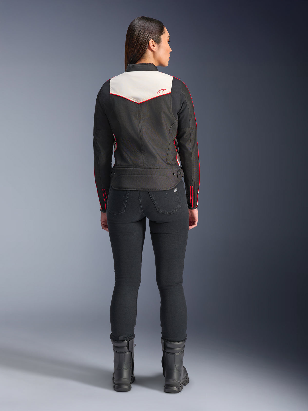 Rückansicht eines weiblichen Models in der Alpinestars Stella T-Dyno WR Jacke in Black Ecru Bright Red, mit einem markanten cremefarbenen Rückenpanel und roten Logo-Akzenten, kombiniert mit einer schwarzen Hose und Stiefeln, eine vintage-inspirierte Textil-Motorradjacke.