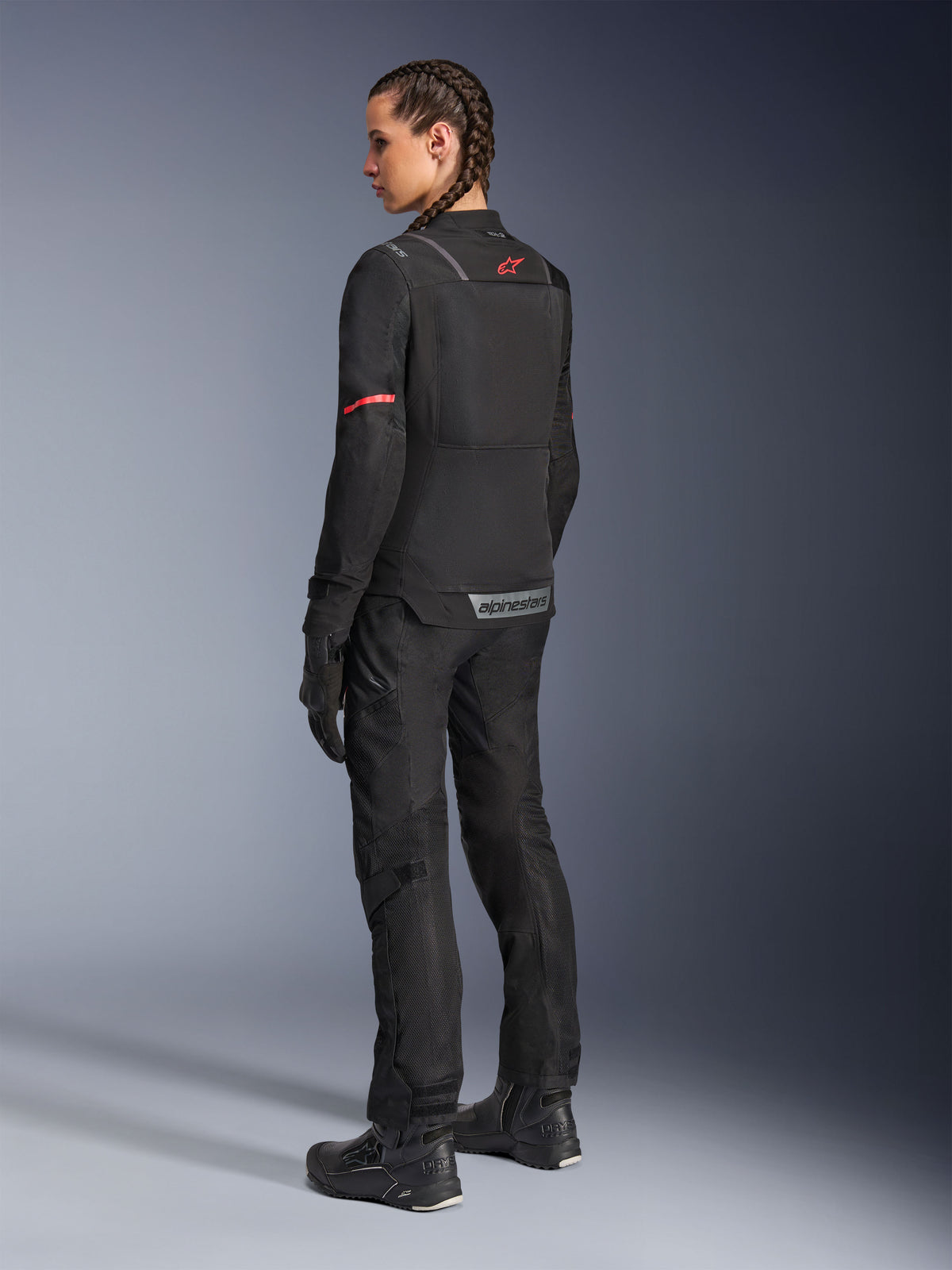 Motorradfahrerin in schwarzer Alpinestars Stella Andes Air Drystar® Hose und dazu passender Stella ST-2 Air Jacke, Rückansicht mit technischer Textilkonstruktion und Mesh-Einsätzen, rotes Stern-Logo auf dem Rücken der Jacke, kombiniert mit schwarzen Motorradstiefeln und Handschuhen.