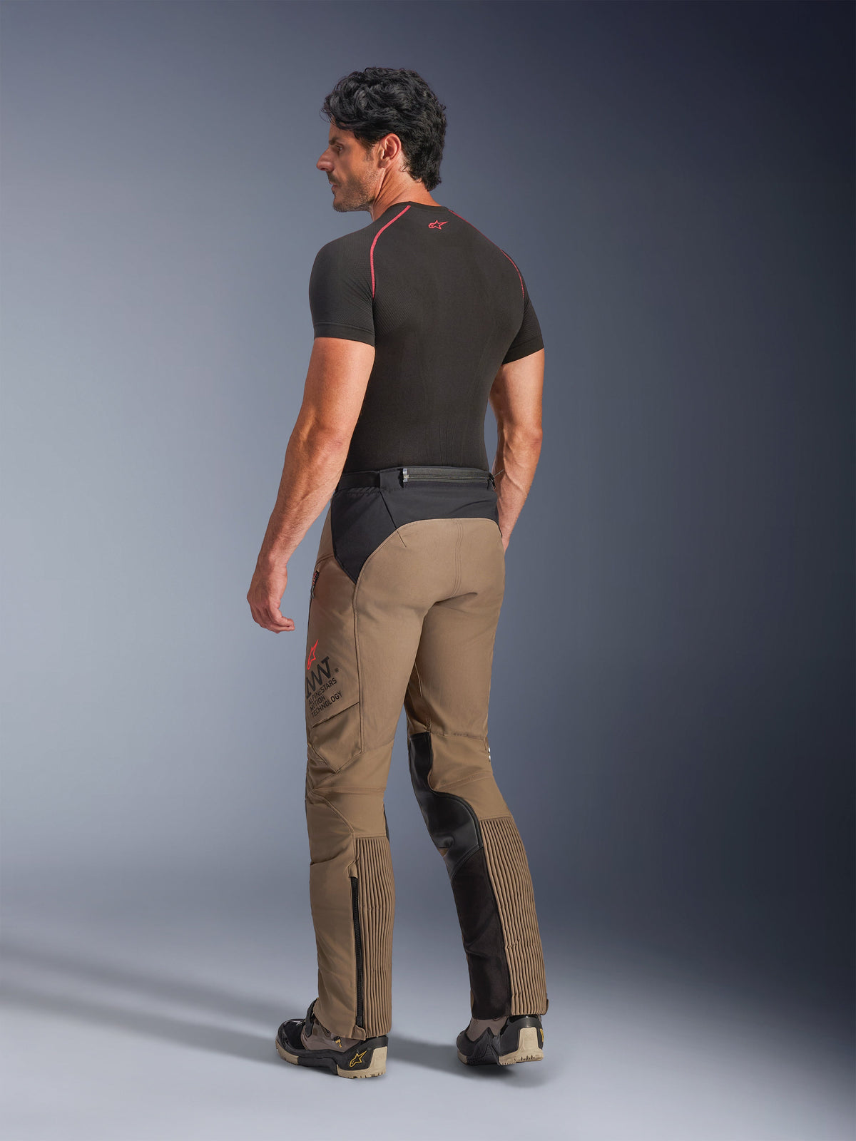 Alpinestars AMT-8 Stretch Drystar® XF Hose in Walnut Black, Adventure-Touring-Motorradhose in der Dreiviertel-Rückansicht, walnussbraunes Textil mit schwarzen Einsätzen an Gesäß und Beininnenseiten, Akkordeon-Stretch-Einsätze an den Waden und AMT-Branding an der Seite, getragen mit einem schwarzen Base Layer.