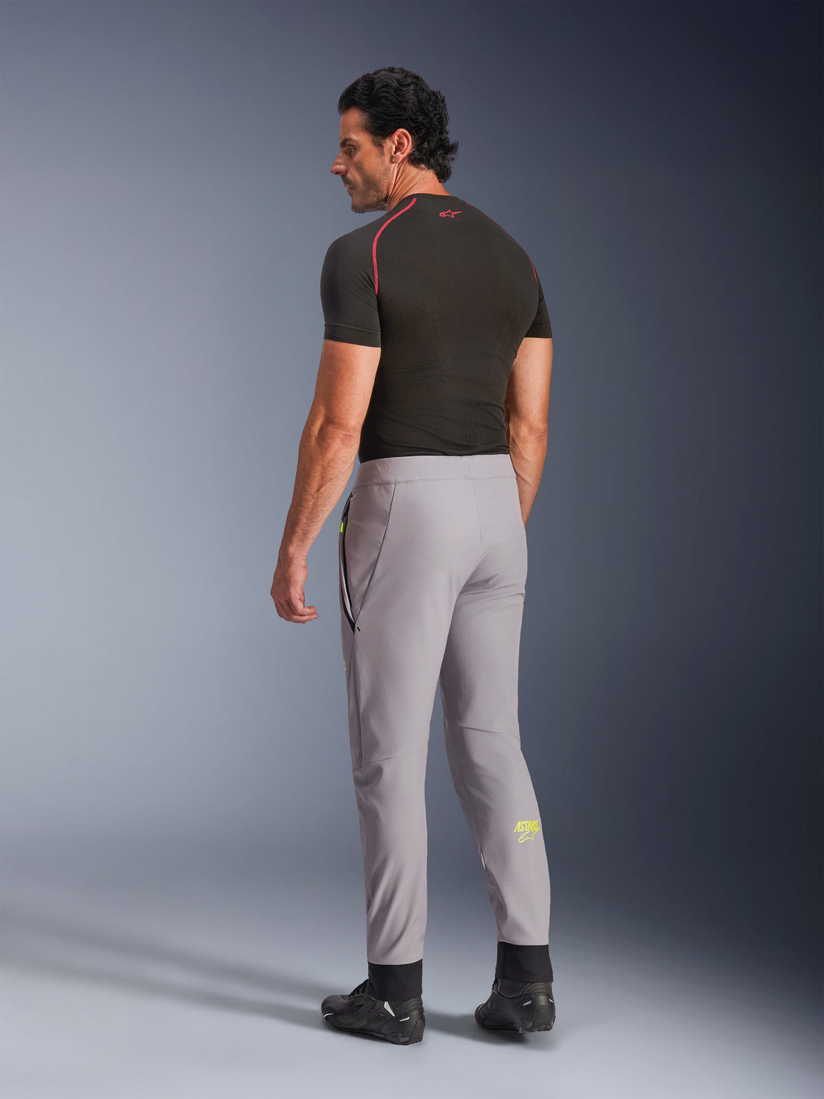 Alpinestars Aeroshell Hose, Mid Gray Yellow Fluo, Rückansicht eines männlichen Modells, das eine Stretch-Softshell-Motorradhose trägt, mit gelbem Astars-Logo auf der linken Wade, kombiniert mit einem schwarzen Base Layer und Motorradschuhen für eine moderne, sportliche Silhouette.