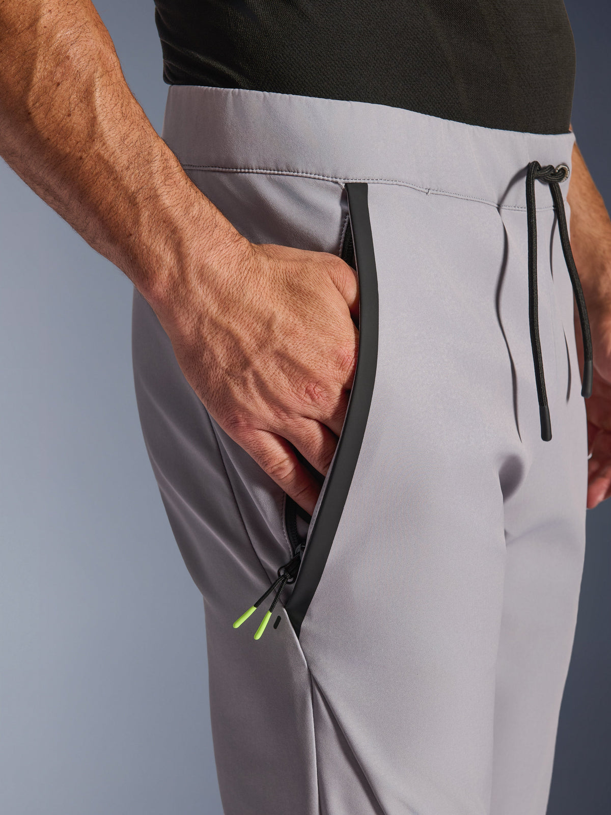 Alpinestars Aeroshell Hose, Mid Gray Yellow Fluo, Nahaufnahme der vorderen Reißverschlusstasche und des Bundes, schwarzer getapter Reißverschluss mit neongelben Enden, elastischer Bund mit schwarzem Kordelzug, Crossover-Sport-Softshell-Motorradhose