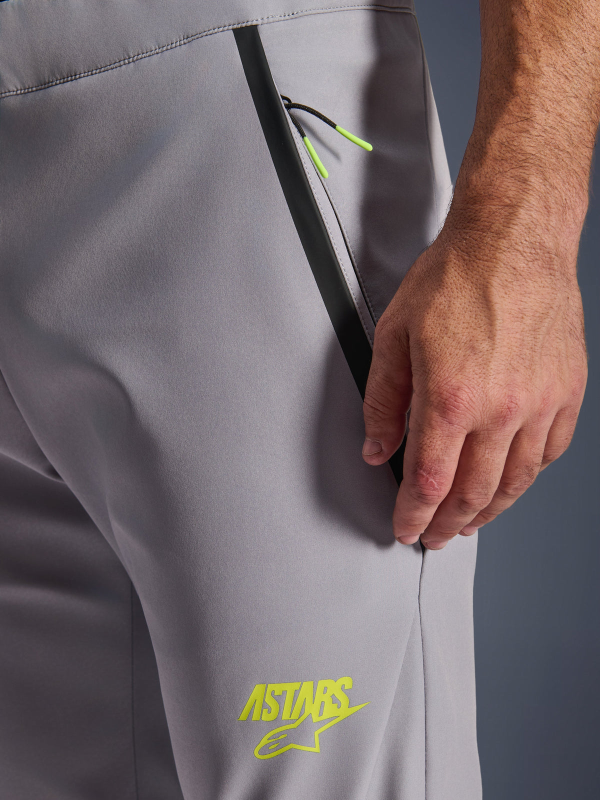 Alpinestars Aeroshell Hose, Mittelgrau mit gelben Fluo-Akzenten, Detailansicht der vorderen Reißverschlusstasche mit schwarzem Tape-Besatz und leuchtend gelben Kordelzügen, gelbes ASTARS-Logo auf dem Oberschenkel, Softshell-Stretchmaterial für das Motorradfahren.