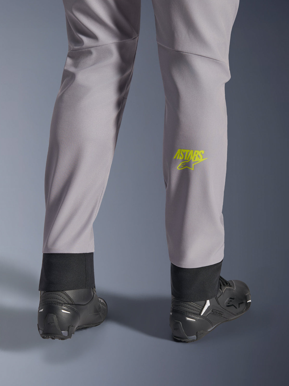 Alpinestars Aeroshell Hose, Mittelgrau mit Yellow Fluo Astars-Logo auf der Wade, Rückansicht mit schwarzen Motorradschuhen, elastischer unterer Saum und moderne, schmal zulaufende Silhouette