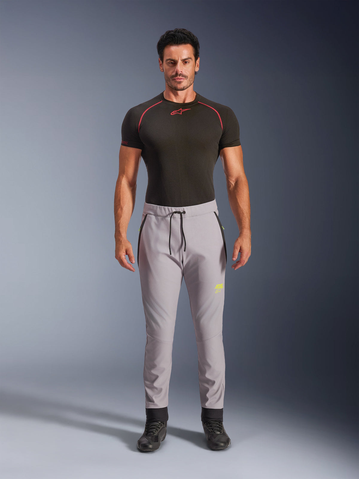 Männliches Model in Alpinestars Aeroshell Hose in Mid Gray und Yellow Fluo, Crossover-Sport-Softshellhose mit elastischem Kordelzug am Bund und Reißverschlusstaschen, kombiniert mit einem schwarzen kurzärmeligen Funktionsshirt, modernes athletisches Design für Straße und Freizeit