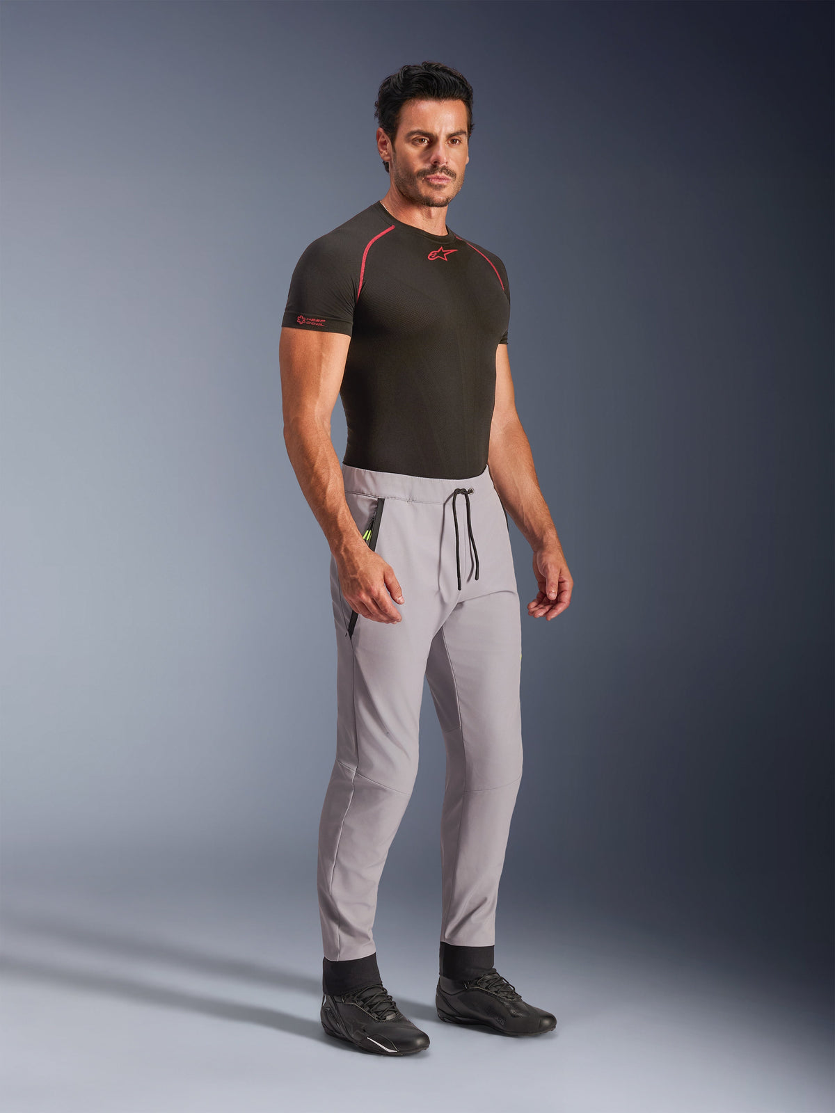 Alpinestars Aeroshell Hose, Mittelgrau mit gelben Fluo-Akzenten, Crossover-Softshell-Hose getragen von einem Model mit schwarzem Baselayer und schwarzen Schuhen, mit moderner, schmal zulaufender Passform, elastischem Bund mit schwarzem Kordelzug und Reißverschlusstaschen.
