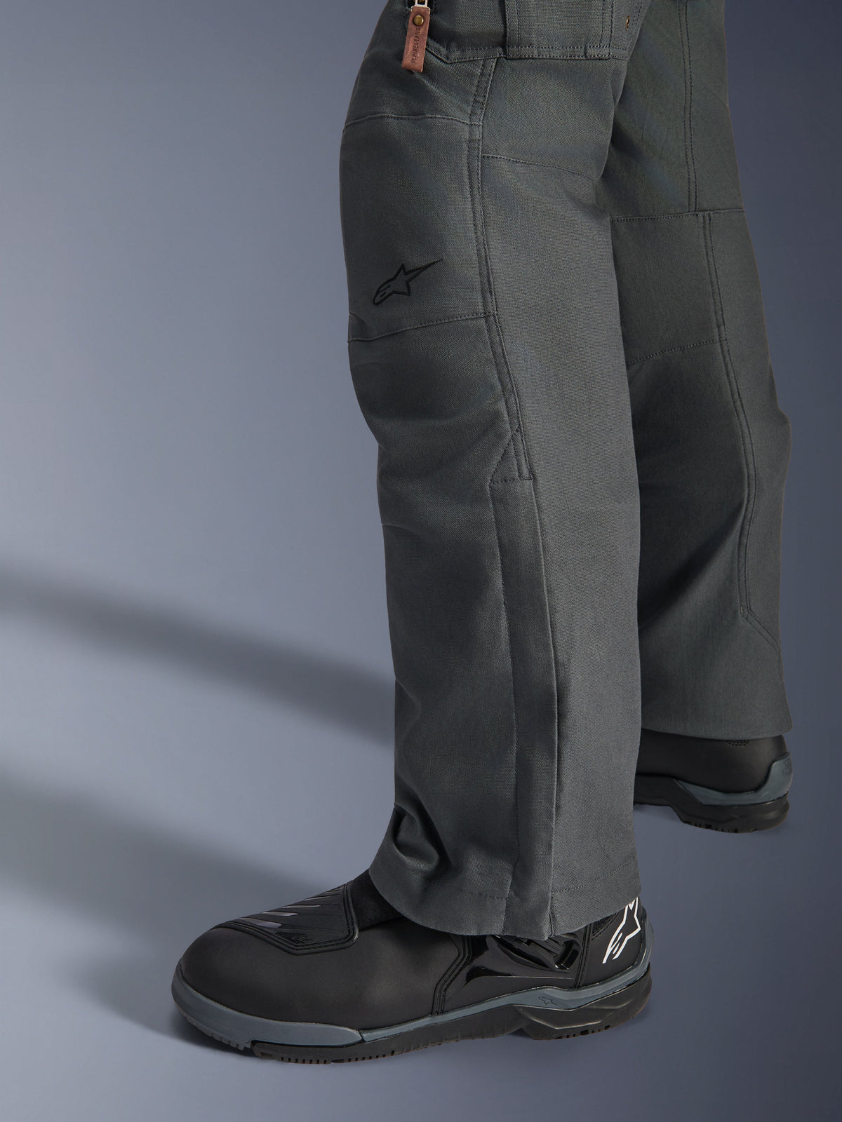 Alpinestars Borrego Drystar® Hose, Abenteuer-Motorradhose in Anthrazitgrau mit gewachster Canvas-Optik, Textilkonstruktion in der Seitenansicht mit schwarzen Motorradstiefeln, robustes Design für Straßen- und Abenteuertouren