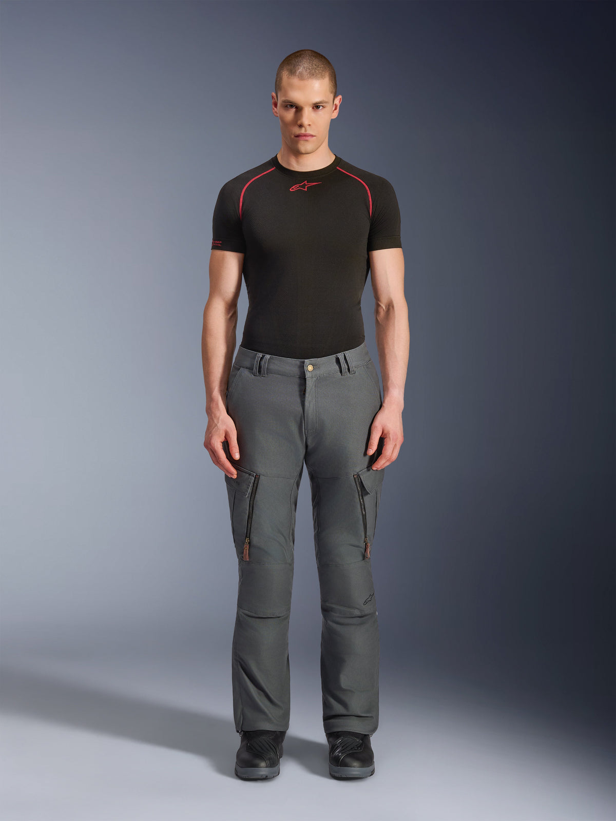 Alpinestars Borrego Drystar® Hose, graue Adventure-Motorradhose, getragen von einem Model mit einem schwarz-roten Funktions-T-Shirt und schwarzen Stiefeln, mit Cargotaschen mit vertikalen Reißverschlüssen, robuster gewachster Canvas-Look, langlebiges Design für Abenteuertouren und Straßenfahrten