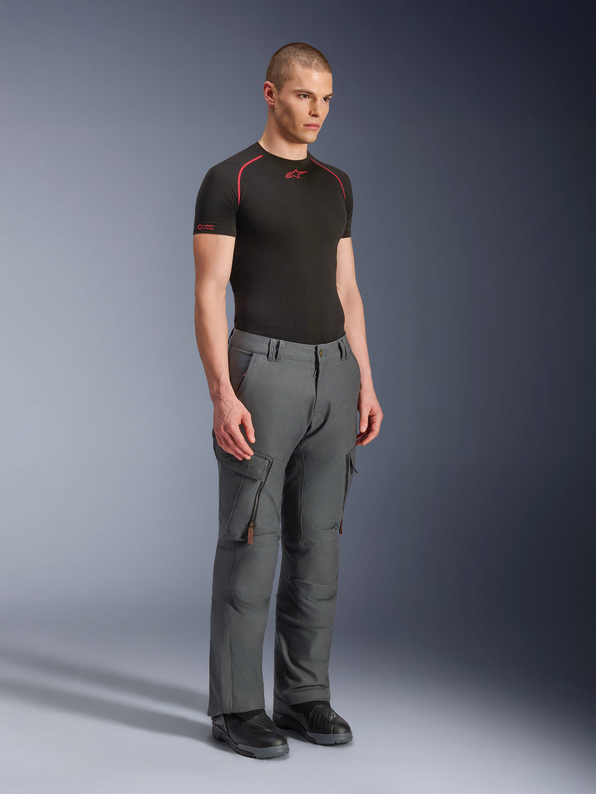 Männliches Model trägt Alpinestars Borrego Drystar® Hose, Adventure-Motorradhose in Grau-Anthrazit, von Arbeitskleidung inspiriertes Design mit Cargo-Taschen und Belüftungsöffnungen mit Reißverschluss, kombiniert mit einem schwarzen kurzärmeligen Base Layer und schwarzen Stiefeln