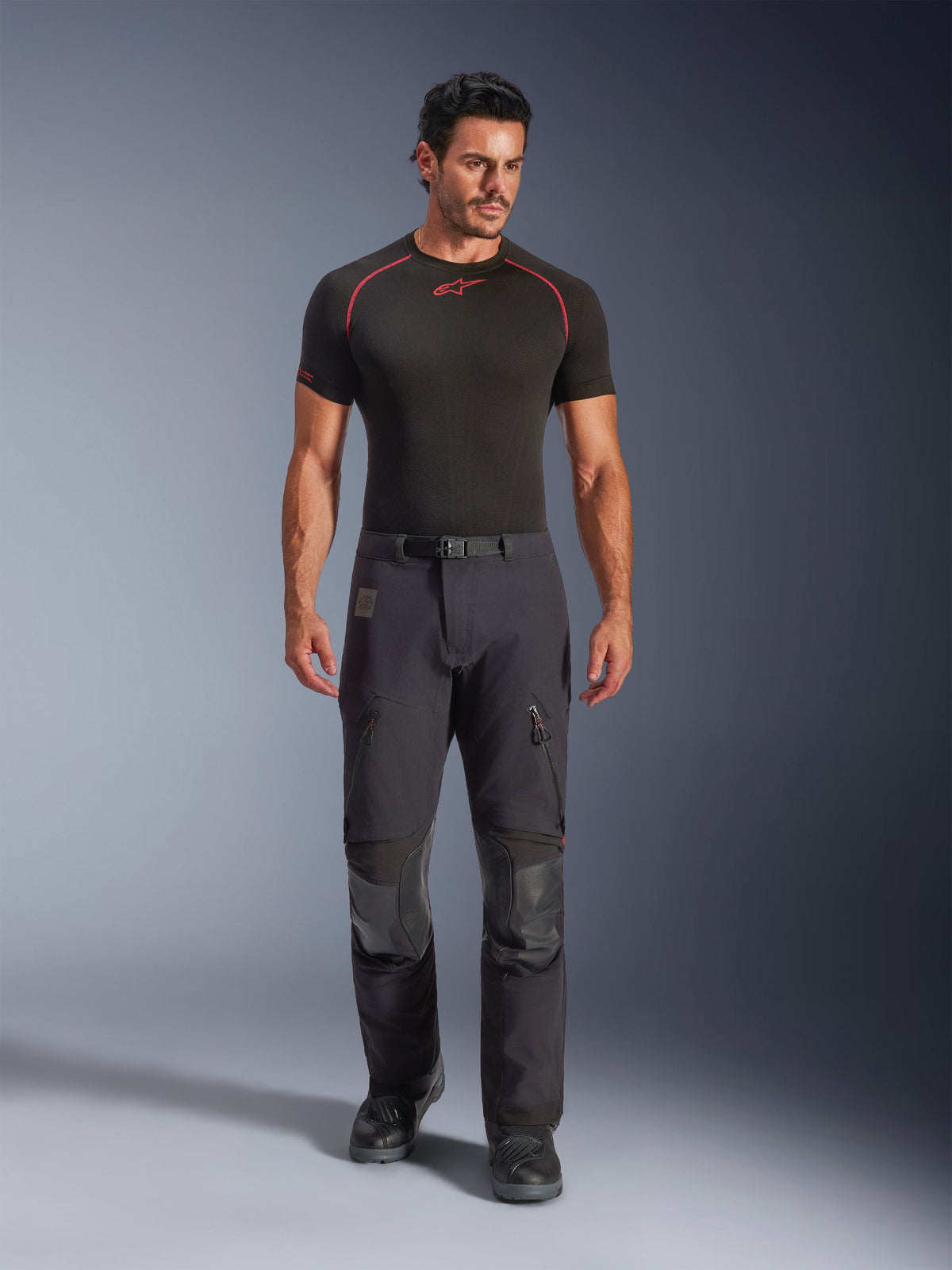 Alpinestars Halo Pro Drystar® XF Laminated Pants in Black Dark Shadow, worn by a model with a black base layer and motorcycle boots, featuring technical zippered thigh vents, cargo pockets, and reinforced knee panels, adventure touring motorcycle gear.