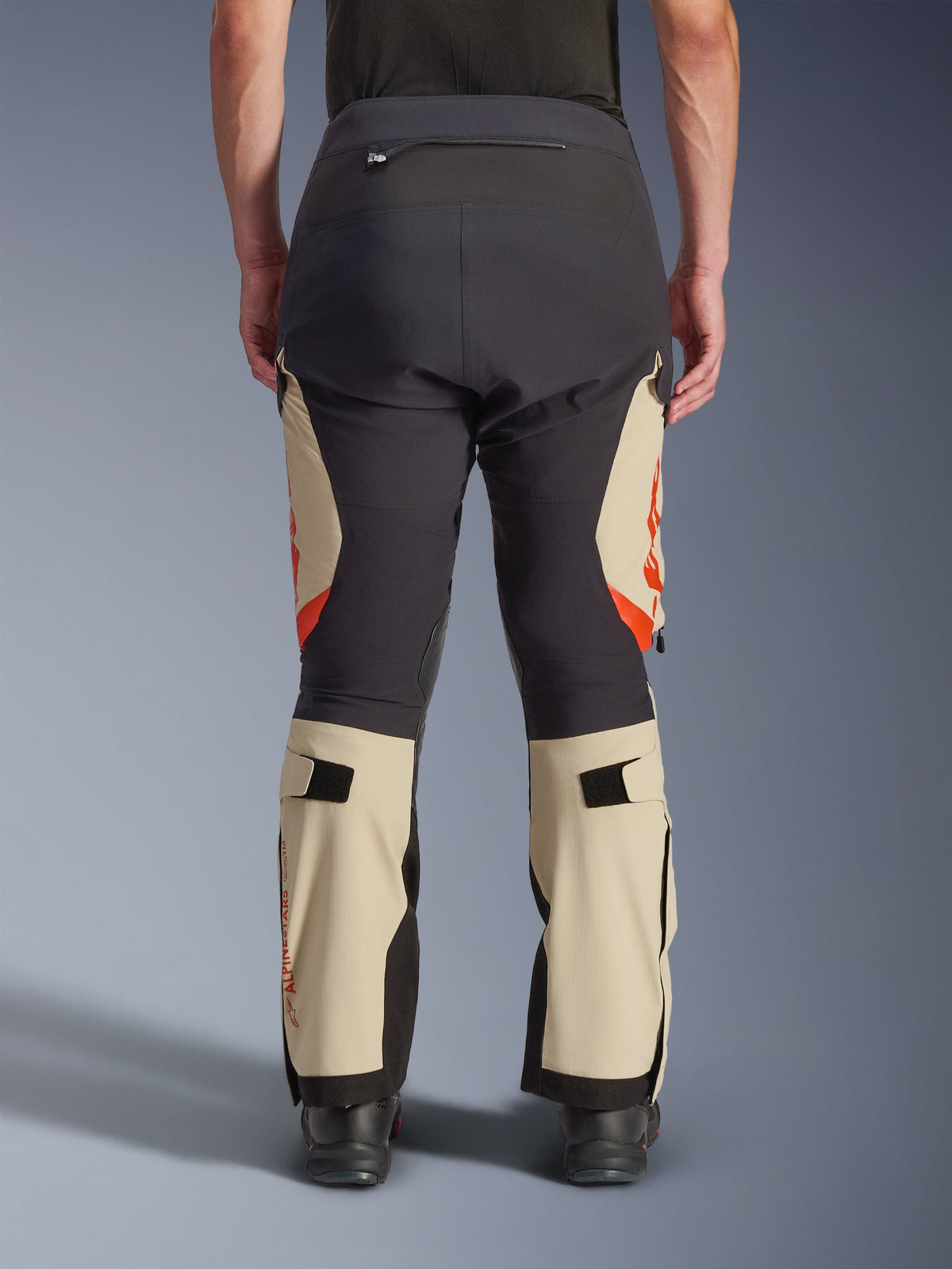 Rückansicht der Alpinestars Halo Pro Drystar® XF Laminierten Hose, Mastic Black mit beigen Einsätzen und orangefarbenen Akzenten, Adventure-Motorradhose mit technischer Textilkonstruktion, getragen mit schwarzen Motorradstiefeln