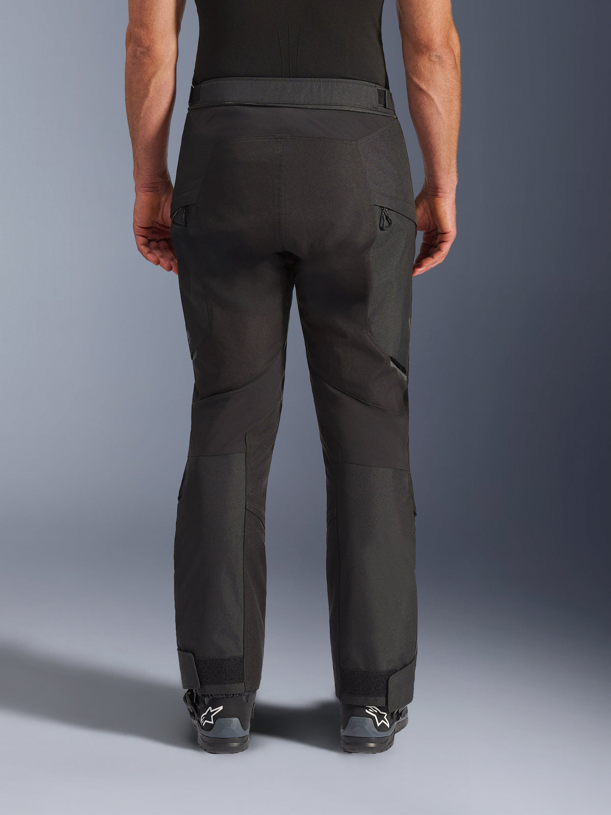 Alpinestars Cusco Drystar® Hose, Motorradhose, Schwarz, Rückansicht getragen von einem Fahrer, mit strukturierten Textilpaneelen, Belüftungsöffnungen mit Reißverschluss an den Oberschenkeln und verstellbaren Knöchelverschlüssen, kombiniert mit schwarzen Alpinestars-Stiefeln.