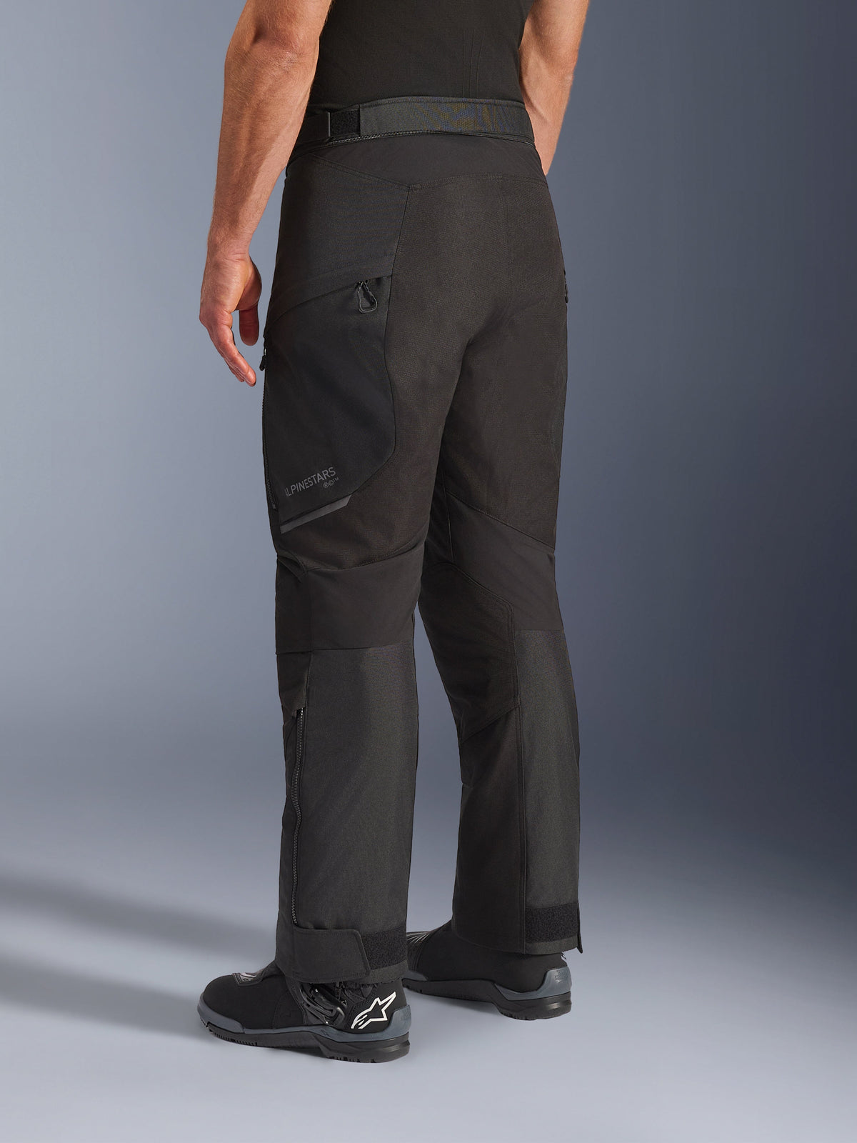 Alpinestars Cusco Drystar® Hose, Motorradhose, Schwarz, Dreiviertel-Rückansicht mit Reißverschlusstasche am Oberschenkel und vorgeformten Paneelen, getragen mit schwarzen Motorradstiefeln, strapazierfähiges Adventure-Touring-Design mit dezentem Branding