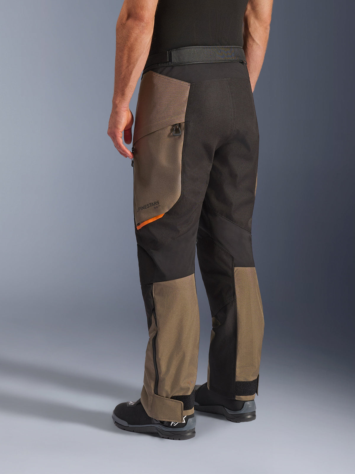 Alpinestars Cusco Drystar® Hose, Adventure-Motorradhose, Walnut Black mit kleinem orangefarbenem Akzent, Dreiviertel-Rückansicht mit Textilkonstruktion und Walnut-Einsätzen, getragen mit schwarzen Motorradstiefeln.