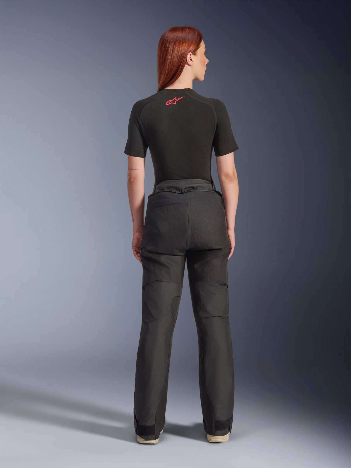 Rückansicht eines Models in der Alpinestars Stella Andes V4 Drystar® Hose in Schwarz, kombiniert mit einem schwarzen, kurzärmeligen Baselayer mit rotem Alpinestars-Logo, Adventure-Touring-Motorradausrüstung mit strapazierfähiger Textilkonstruktion.