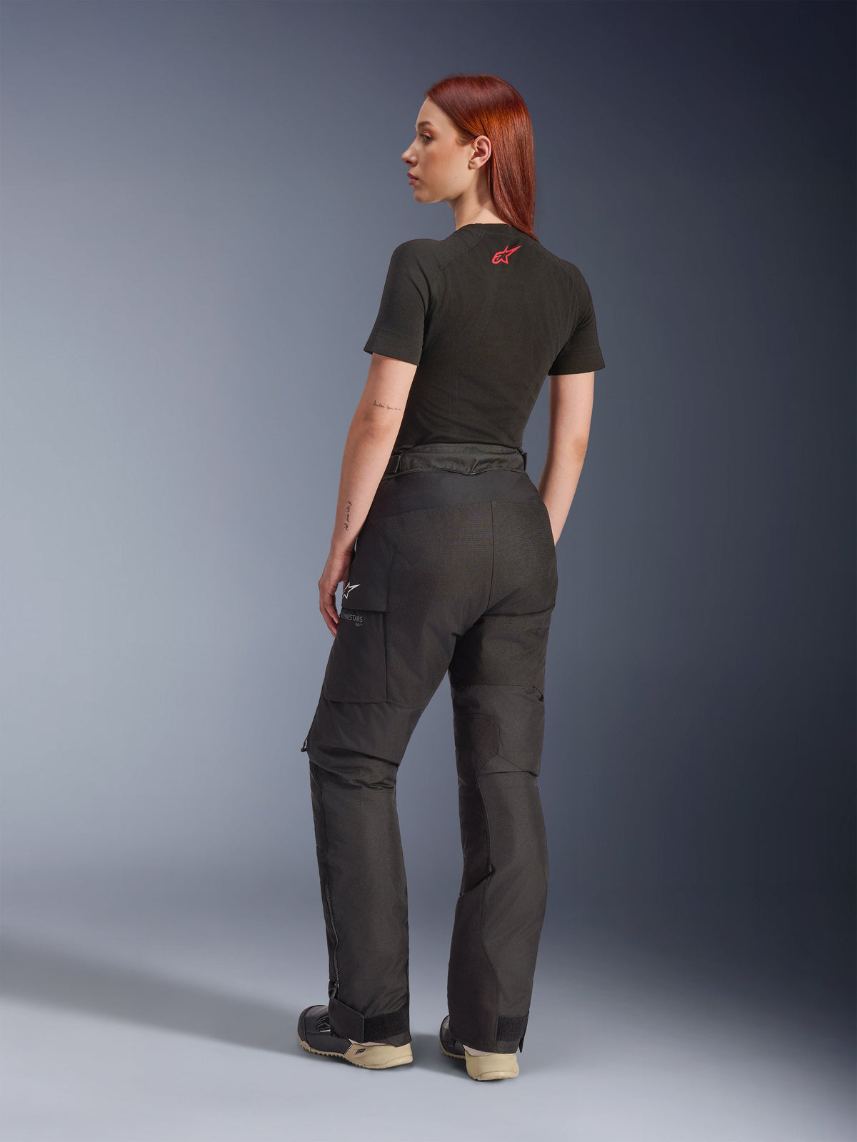 Alpinestars Stella Andes V4 Drystar® Hose, Schwarz, Adventure-Motorradhose in der Seiten- und Rückansicht an einem Model, kombiniert mit einem schwarzen T-Shirt und Stiefeln, mit seitlichen Cargo-Taschen mit weißem Logo-Branding und strapazierfähiger Textilkonstruktion.