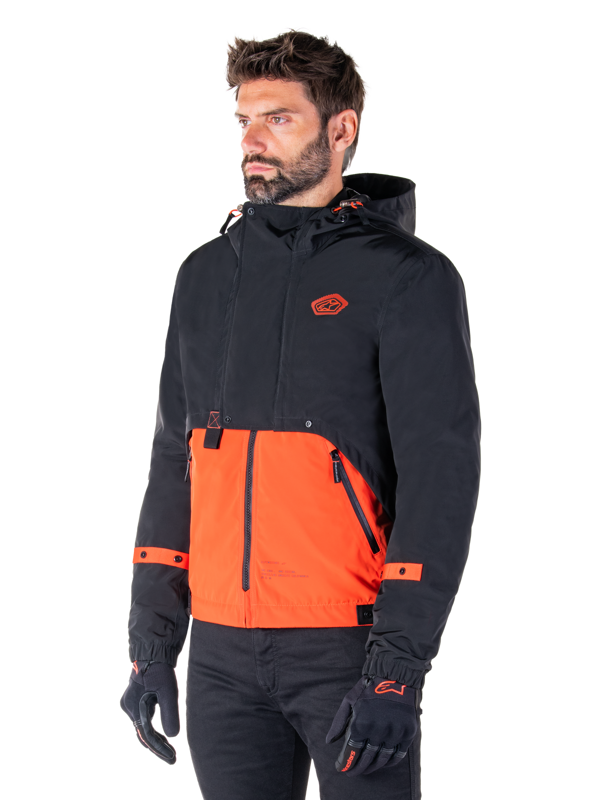 Man wearing the Alpinestars Mootant Waterproof Parka in Spicy Orange and Black, a modular motorcycle jacket with a black hooded upper layer and bright orange lower section, styled with black riding gloves and pants.