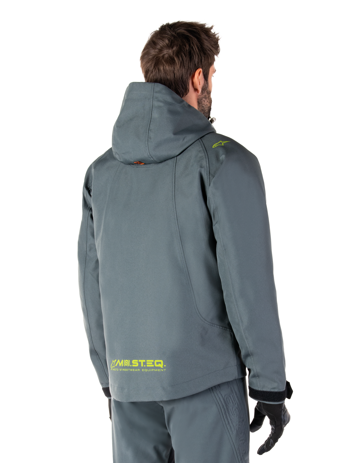 Alpinestars Morush Wasserdichter Parka in Urban Gray, Rückansicht, mit gelbem MO.ST.EQ. Branding am unteren Rücken und an der Schulter, minimalistisches Kapuzendesign, getragen mit passender grauer Motorradhose und schwarzen Handschuhen, urbane Motorrad-Streetwear.