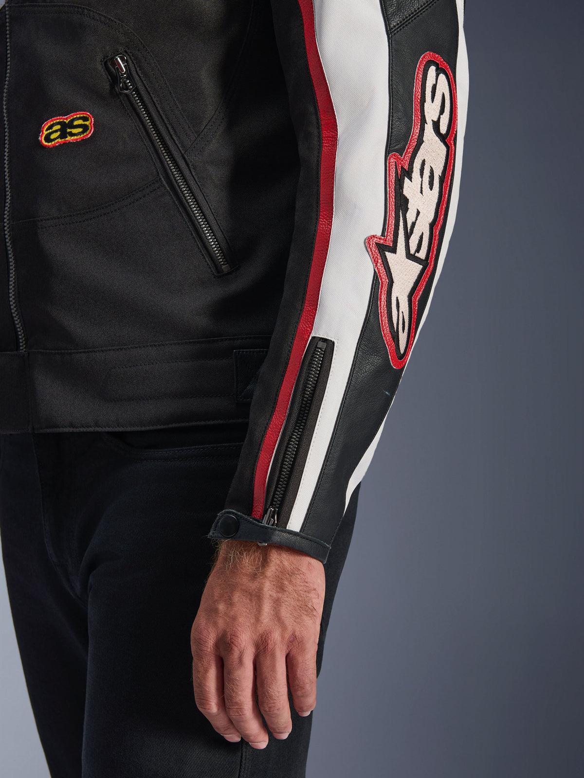 Alpinestars Axton Jacke, Nahaufnahme von Ärmel und Torso, Schwarz mit Off-White und leuchtend roten Akzenten, großes besticktes Alpinestars-Logo am Arm, vertikale Rennstreifen, Ärmelbündchen mit Reißverschluss und Druckknopf, robuste Textilkonstruktion