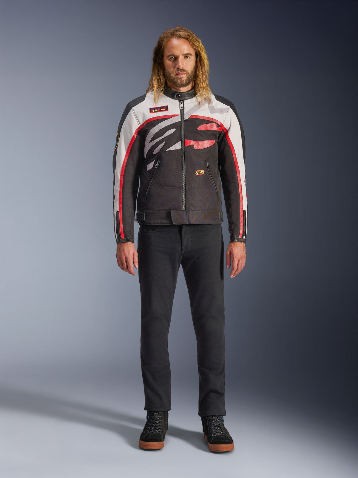 Alpinestars Axton Jacke, Motorradjacke, Schwarz, Off-White und Leuchtendes Rot, getragen von einem Model mit schwarzer Hose und Sneakern, mit einem markanten, umlaufenden Logo und einem vom Vintage-Rennsport inspirierten Design