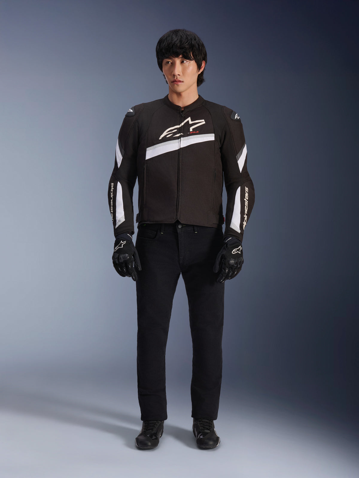 Alpinestars T-GP Plus R V4 Jacke, Schwarz und Weiß, Sport-Textilmotorradjacke, getragen von einem Modell mit schwarzer Hose und Handschuhen, mit markantem weißen Alpinestars-Logo auf der Brust und den Ärmeln, schlankes, vom Rennsport inspiriertes Design
