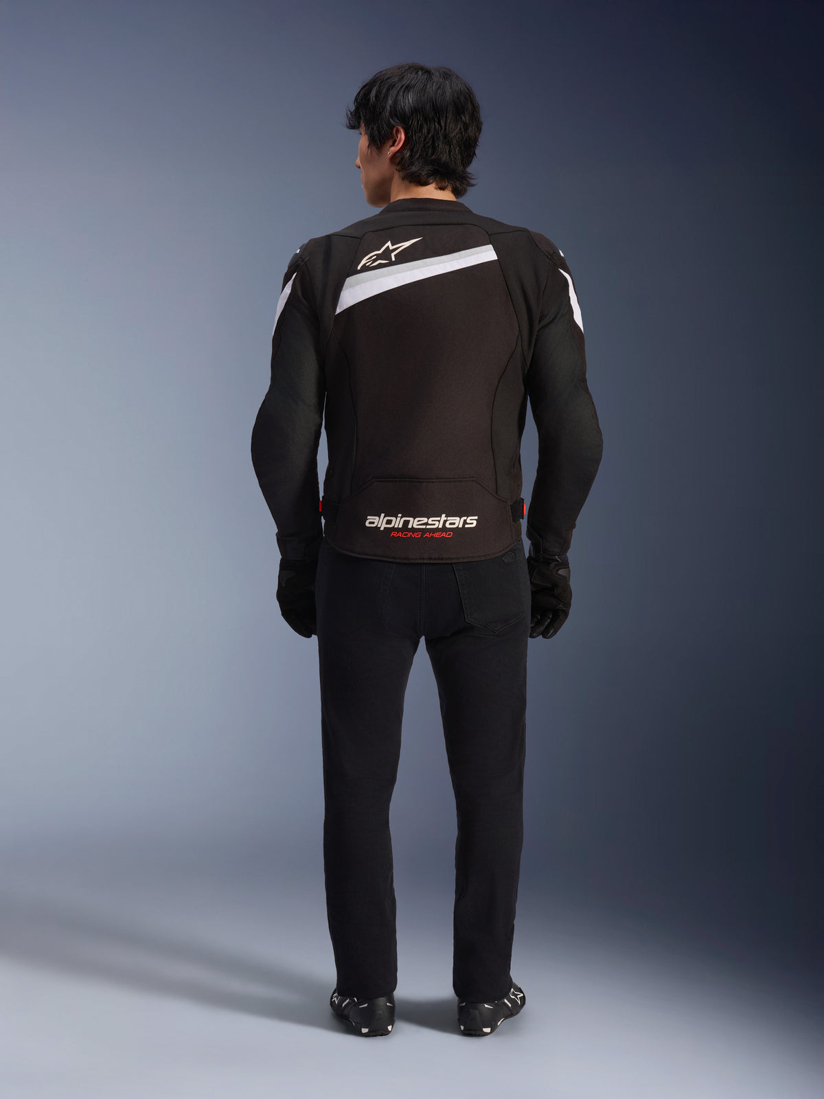 Alpinestars T-GP Plus R V4 Jacke, sportliche Textil-Motorradjacke in Schwarz und Weiß, Rückansicht mit weißem Alpinestars-Logo und „RACING AHEAD“-Branding, getragen mit schwarzer Hose und Stiefeln, stromlinienförmiges Design für sportliches Fahren.