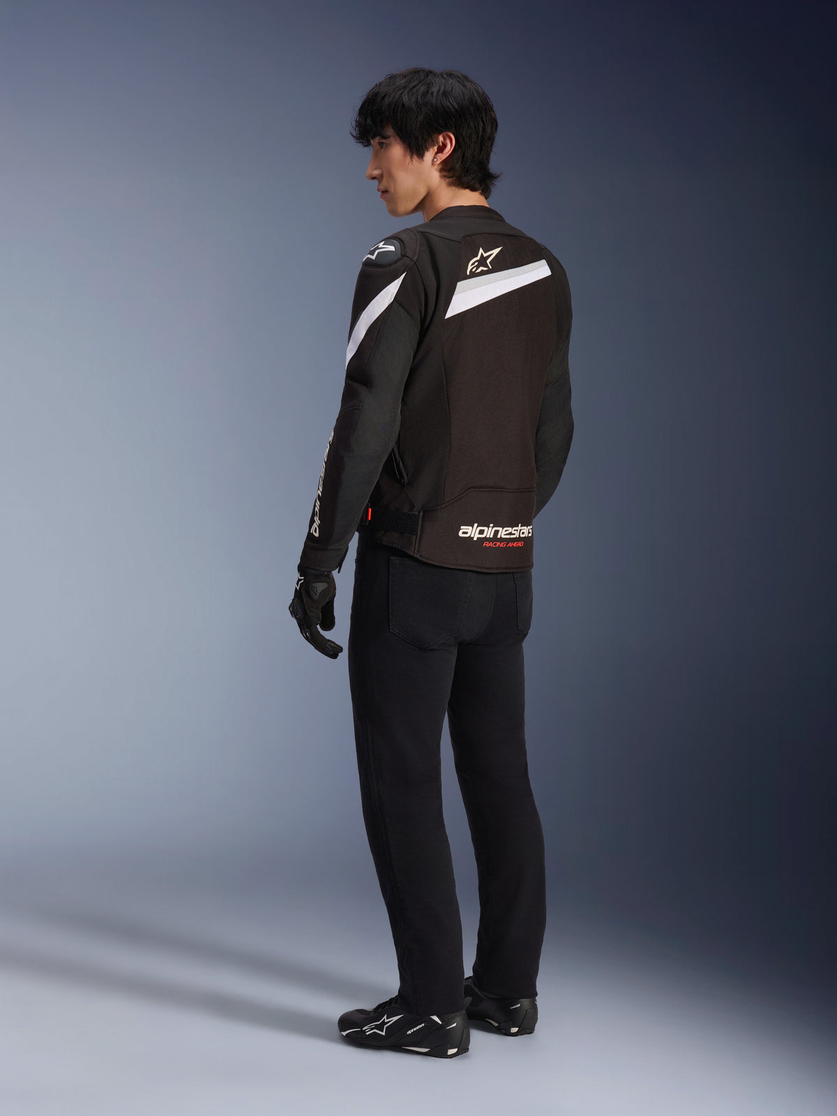 Männlicher Fahrer in der Alpinestars T-GP Plus R V4 Jacke in Schwarz und Weiß, Rückansicht mit weißen diagonalen Akzenten und Branding am unteren Rücken, kombiniert mit schwarzer Motorradhose, Handschuhen und technischen Stiefeln, sportlich geschnittene Textil-Motorradausrüstung